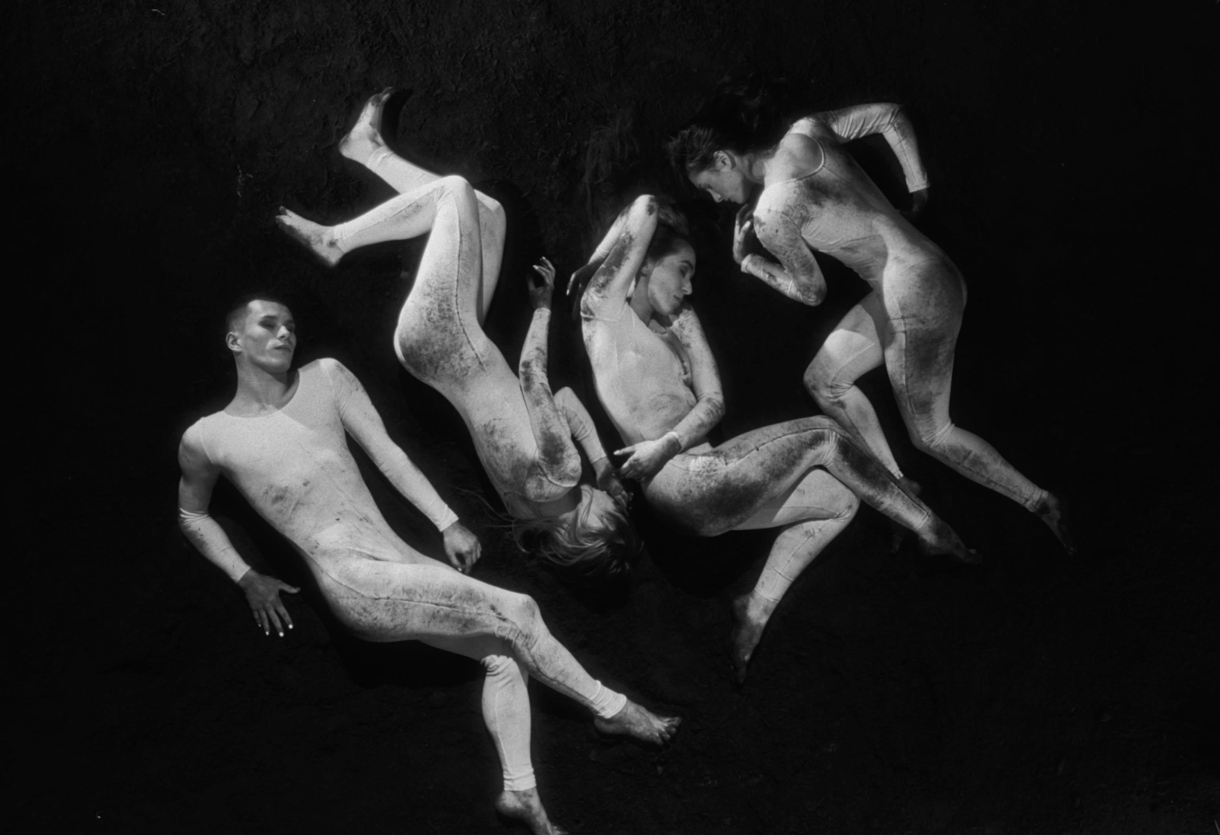 Four women in dance poses lying on a dark floor, dressed in white bodysuits, with a contrasty black-and-white filter.