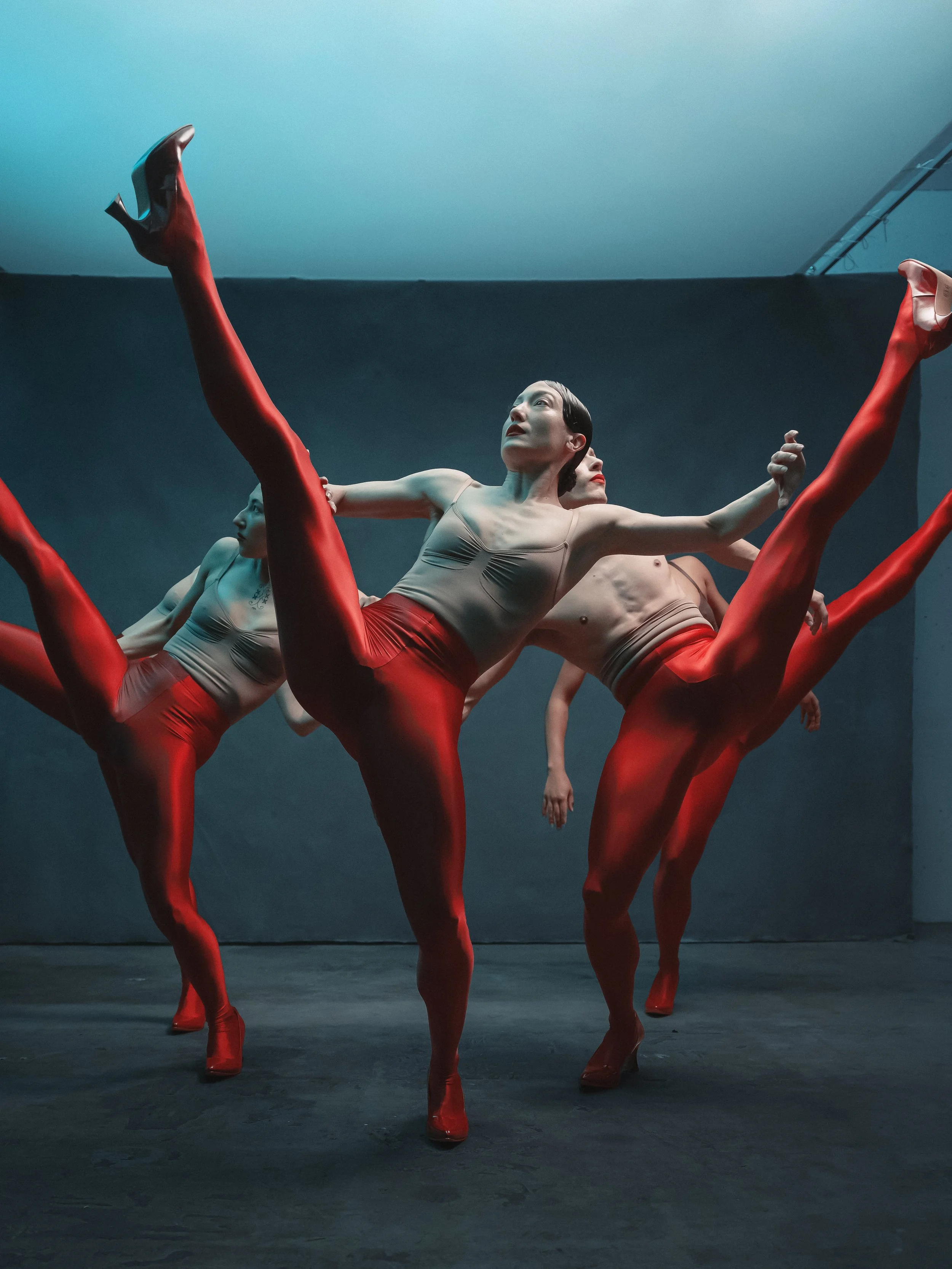 A group of ballerinas practicing on stage, wearing red leggings, beige leotards, and high-heeled shoes, performing in a synchronized dance move.