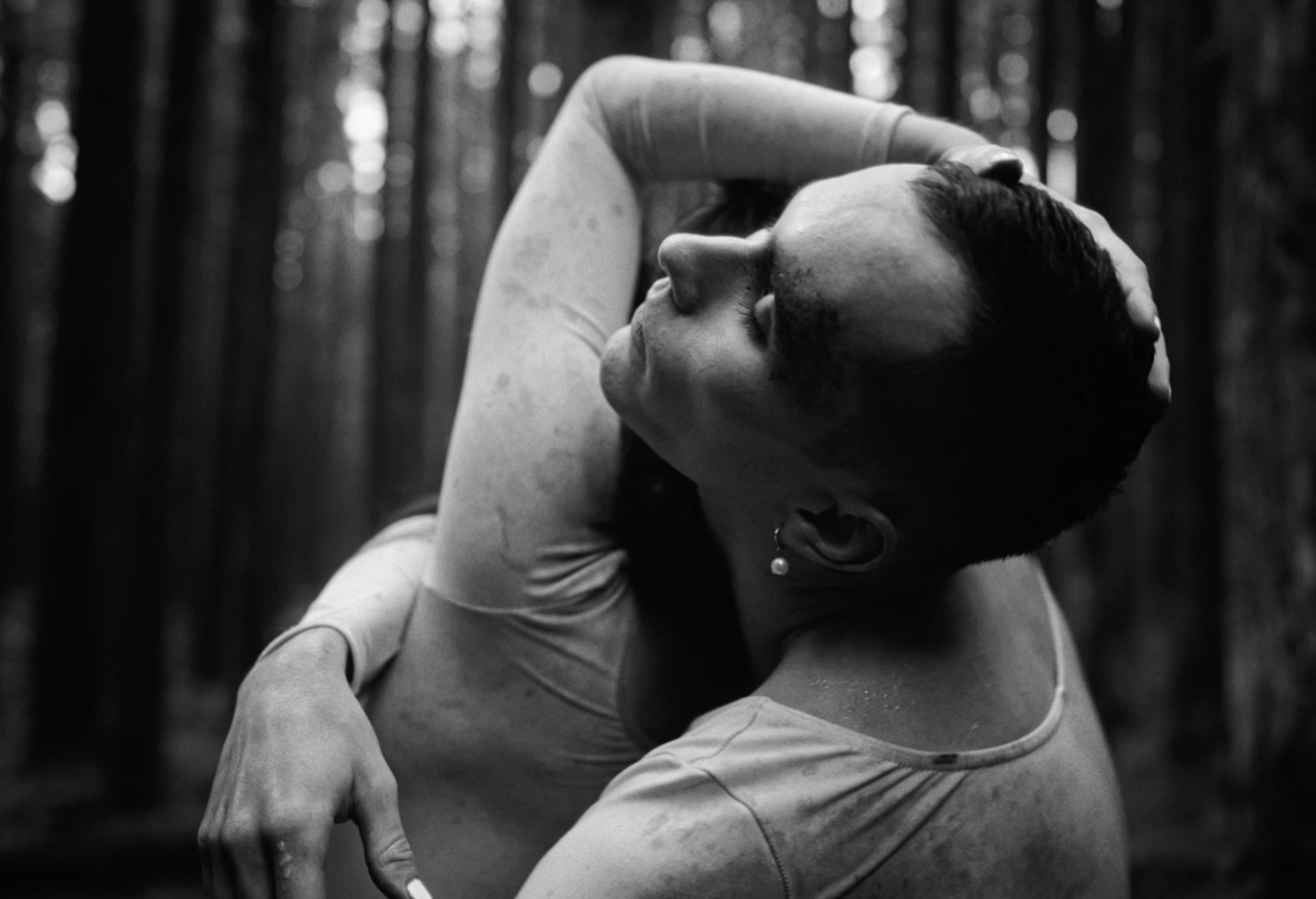 A woman with short hair and earrings in a forest, holding her head with one arm while squatting.