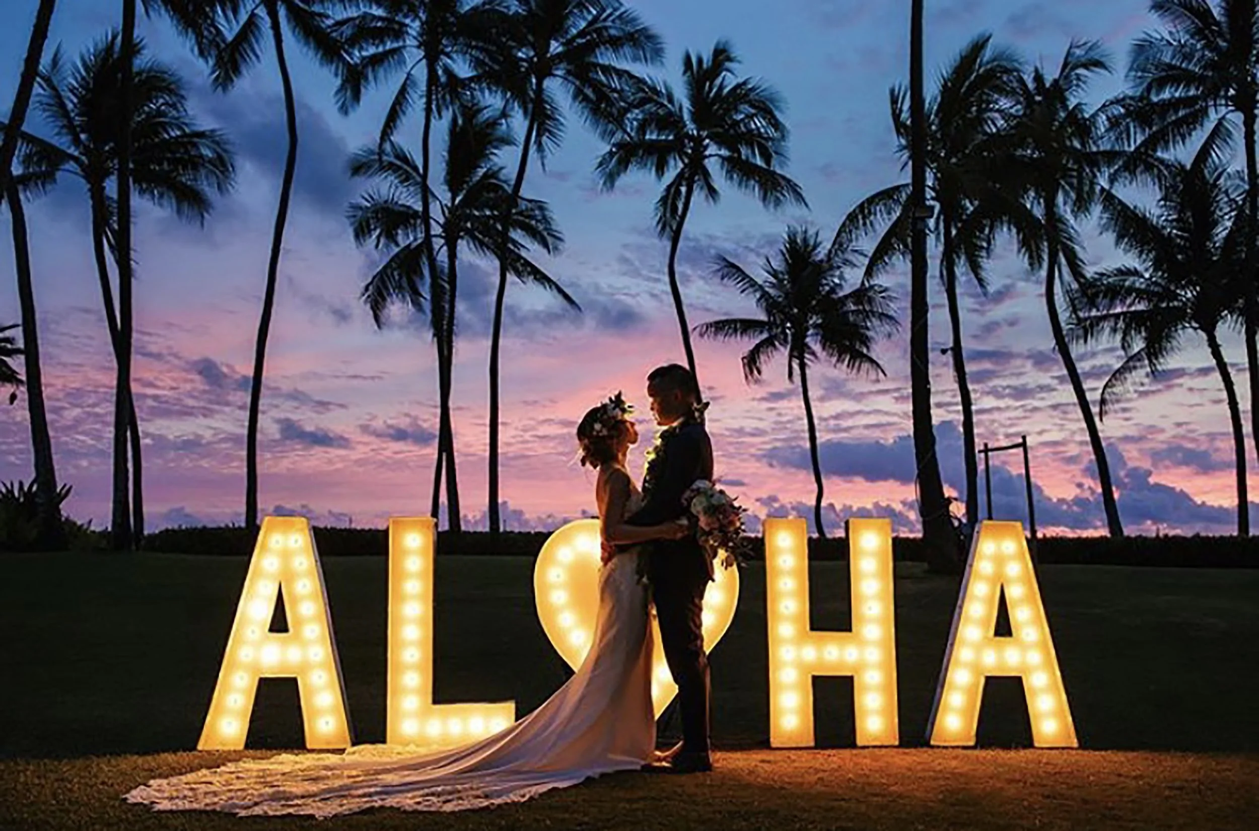 Romantic wedding couple posing by illuminated ALOHA marquee with heart symbol, palm trees, and vibrant Hawaiian sunset sky.