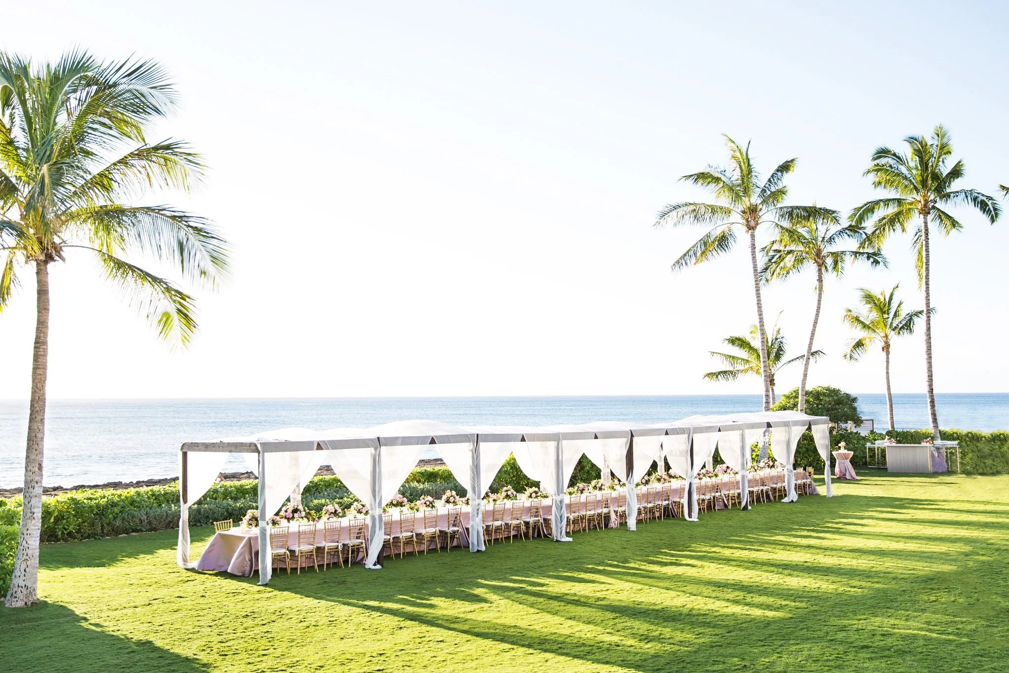 Elegant outdoor wedding reception setup with canopy, floral centerpieces, gold chairs, palm trees, and ocean view in tropical Hawaii.