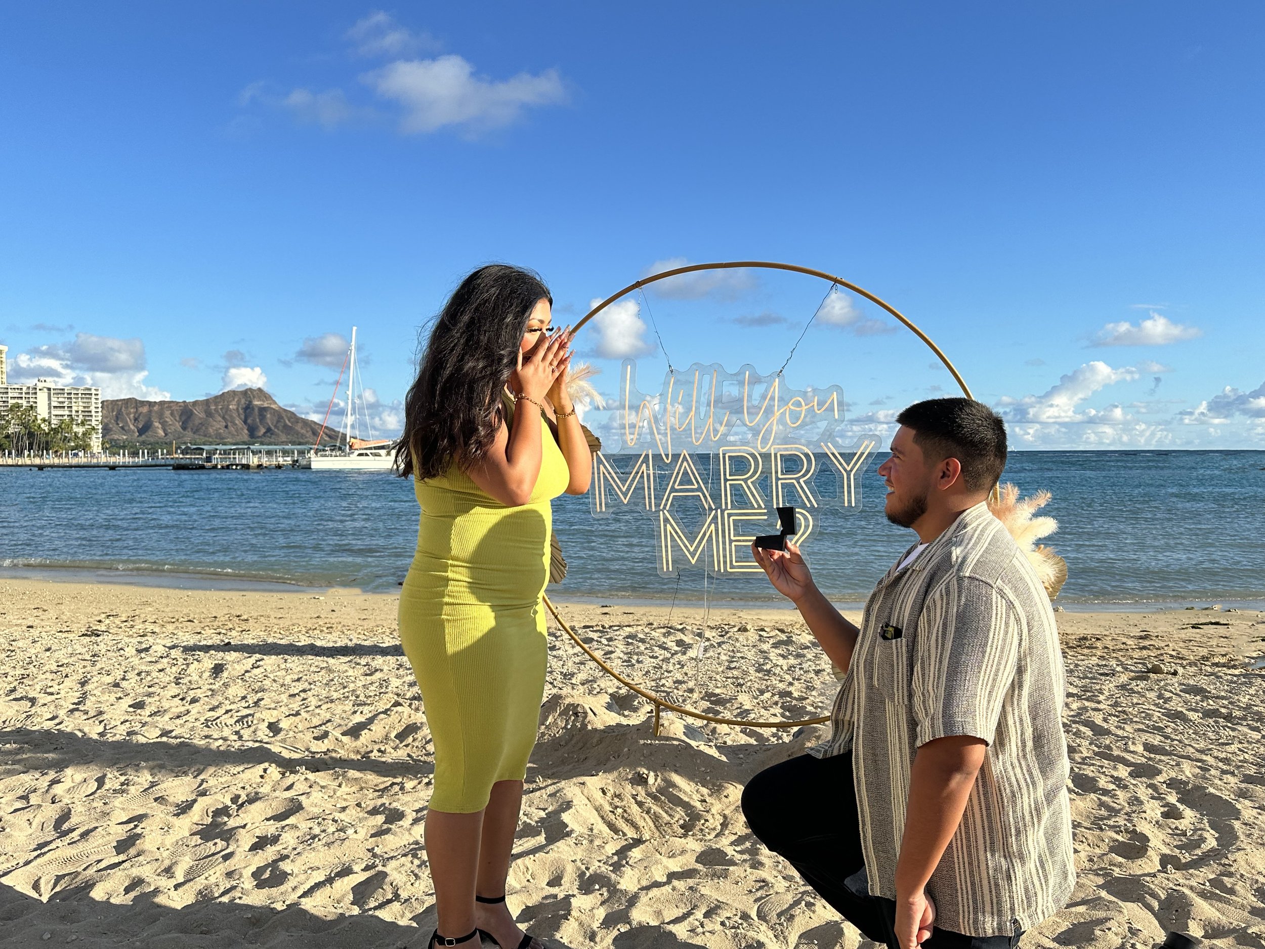 Beach marriage proposal with man kneeling, ring box, illuminated 'Will You Marry Me?' sign, ocean view, boats, and tropical mountain backdrop.