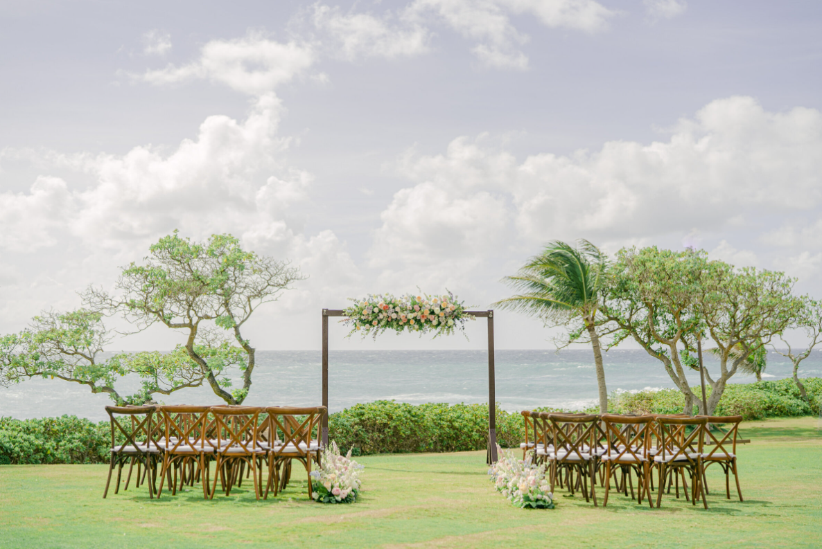Beachside wedding ceremony with wooden arch and pastel floral arrangements, aisle lined with chairs, overlooking ocean and tropical scenery.