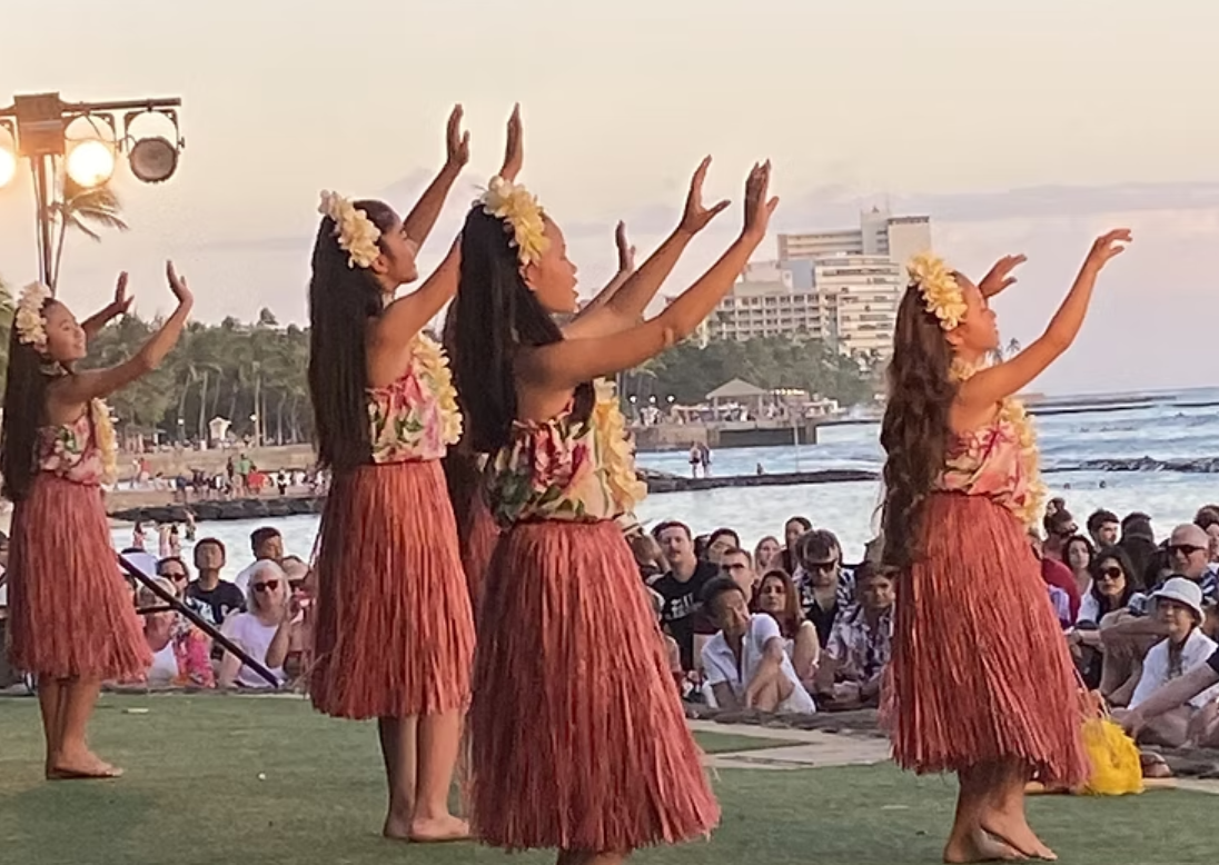Group of dancers performing traditional Hawaiian hula in red grass skirts and floral leis on a beachside stage at sunset, with ocean and palm trees in the background.