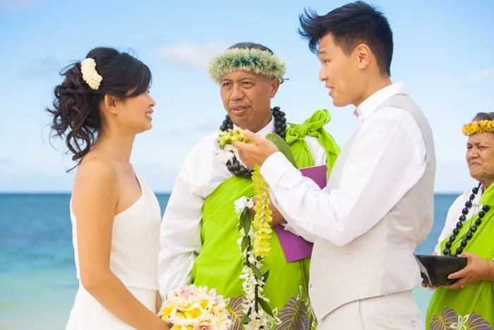 Hawaiian beach wedding ceremony with couple exchanging leis, guided by traditional officiants in green robes, ocean backdrop.