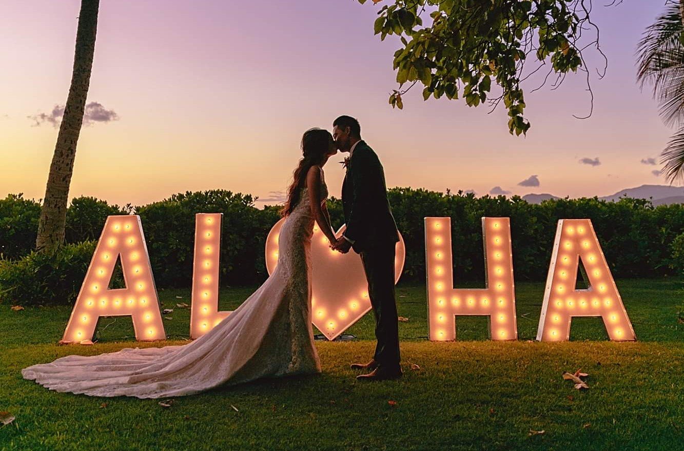Wedding couple holding hands in front of illuminated ALOHA letters at sunset, tropical outdoor ceremony with palm trees and mountains in the background.