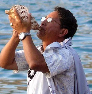 Person performing Hawaiian conch shell ceremony near water, wearing traditional attire, symbolizing cultural ritual.