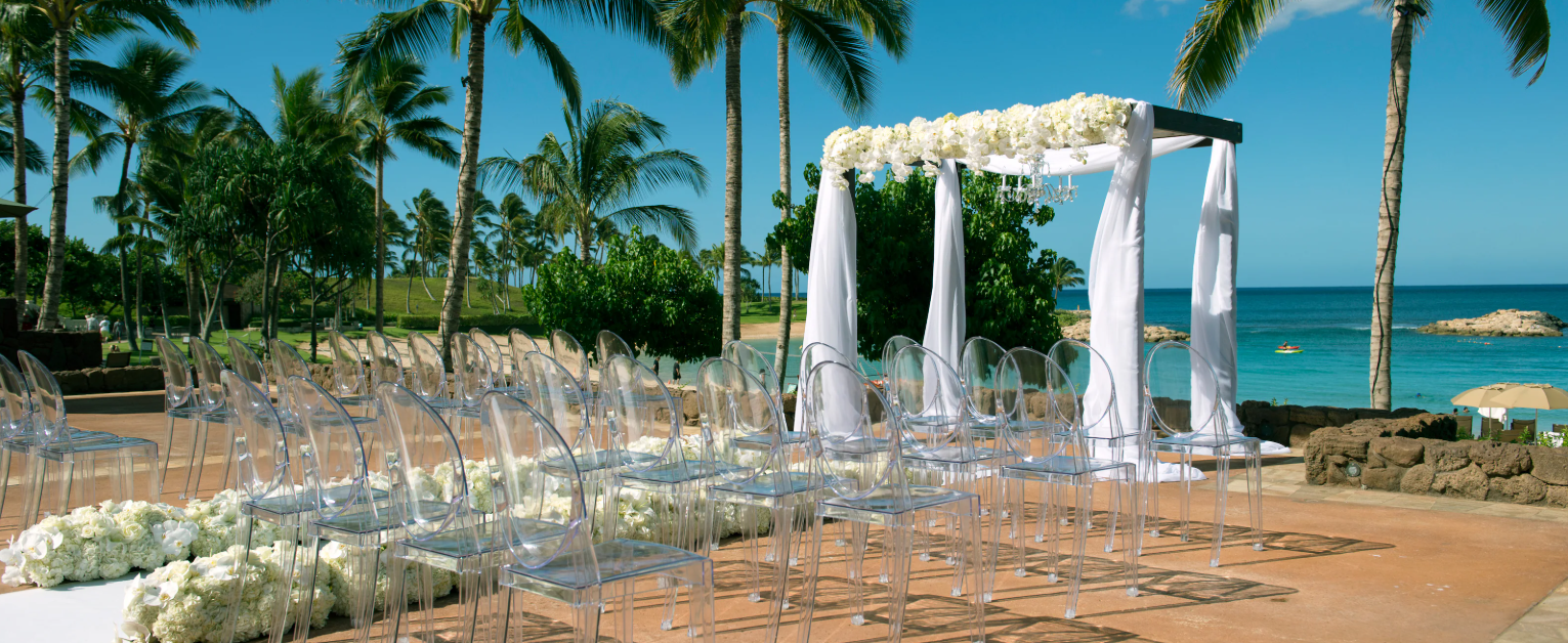 Beachfront wedding ceremony with transparent chairs, white floral arch, palm trees, turquoise ocean, and tropical scenery in Hawaii.