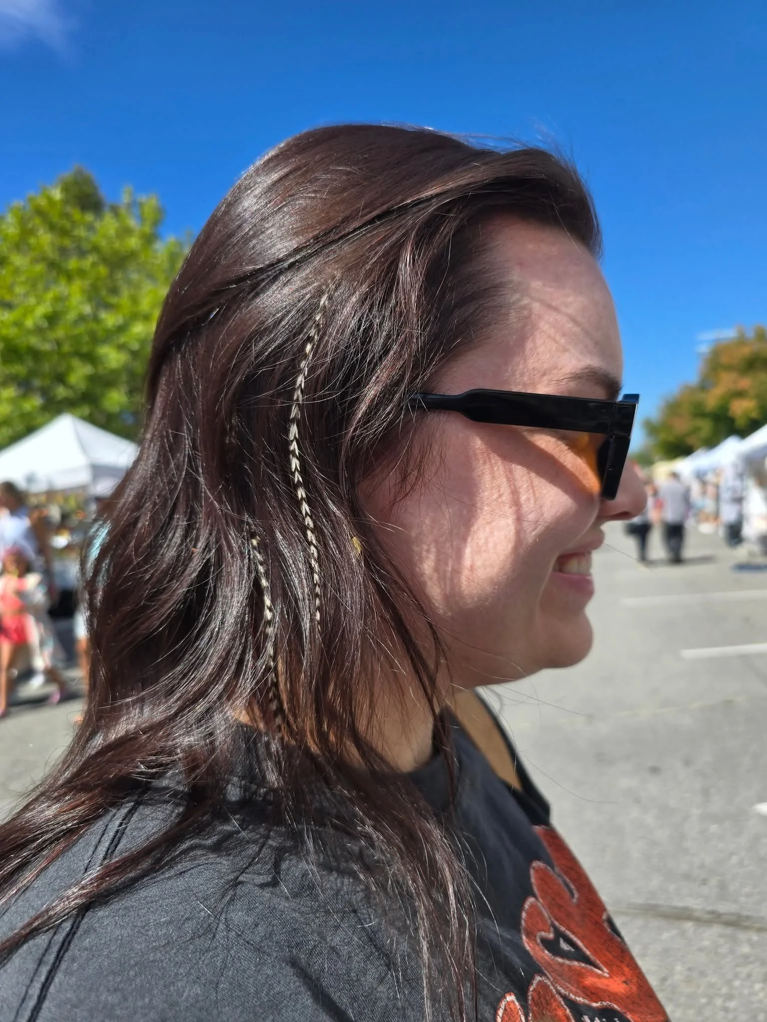 Side profile of a smiling woman with long brown hair and orange-tinted sunglasses, at an outdoor event on a sunny day with tents and trees in the background.
