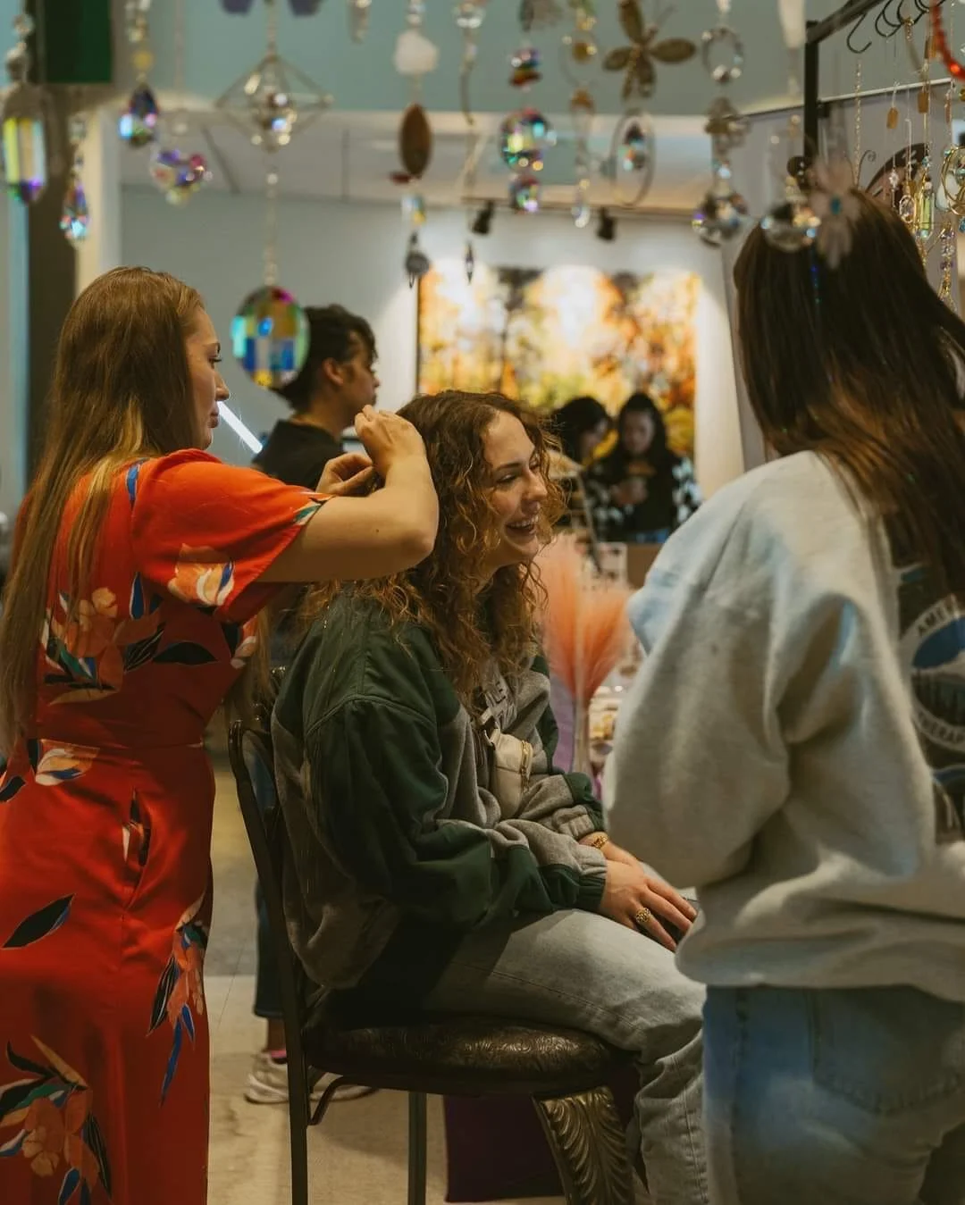 Three women are having hair tinsel applied, with vibrant suncatcher decorations hanging in the background.