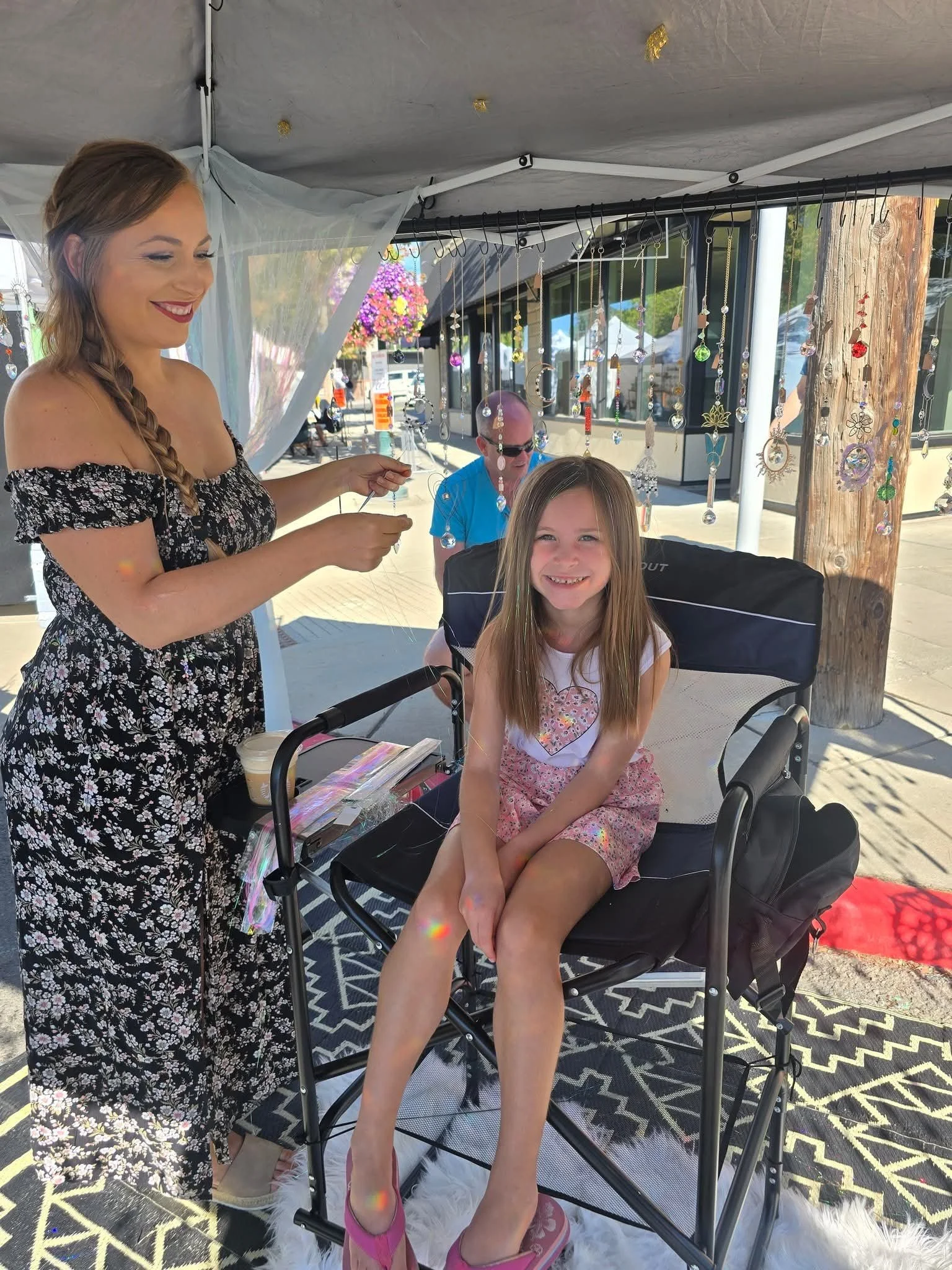 A woman with a braid is smiling and holding hair TINSLE in front of a young girl sitting in a chair, both under a tent with hanging suncatcher decorations, outdoors with buildings and flower baskets in the background.