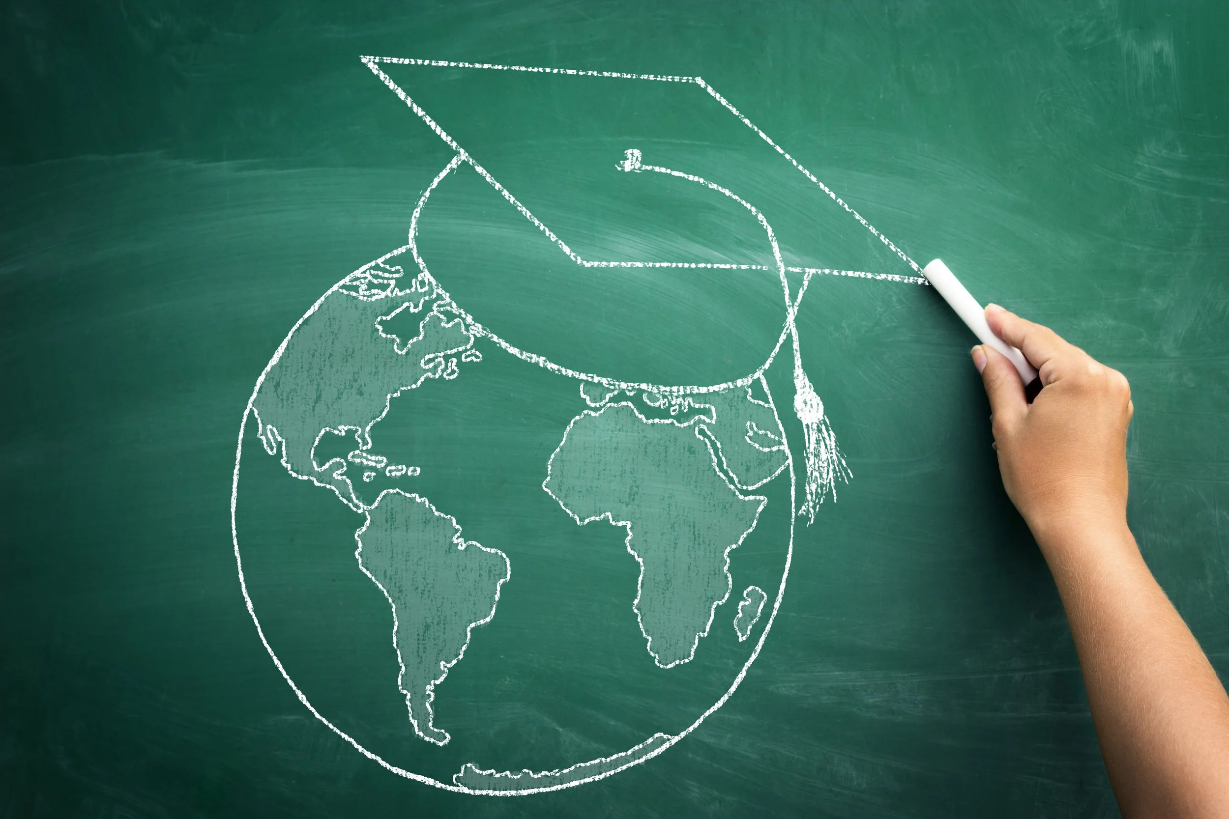 A hand drawing a chalk outline of a graduation cap on top of a world map drawn on a green chalkboard.