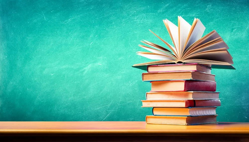 Stack of hardcover books with an open book on top, placed on a wooden surface against a teal background.