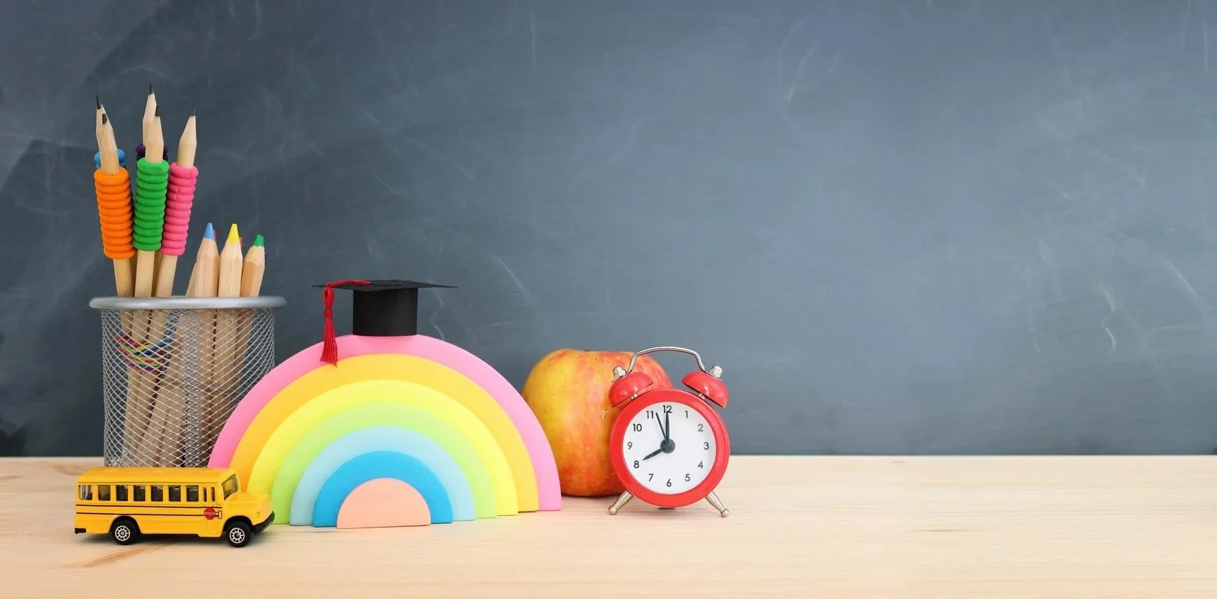 School supplies on a wooden desk with a blackboard in background. Items include a metal mesh pencil holder with colorful pens, a rainbow-shaped organizer with a graduation cap, an apple, a red alarm clock, and a small yellow school bus toy.