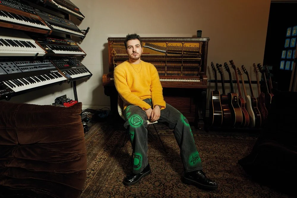 A young man with dark hair and a beard wearing a yellow fuzzy sweater and gray pants with green glow-in-the-dark patches, sitting in a music studio surrounded by musical instruments. Behind him are several electric and acoustic guitars, a vintage upright piano, and multiple keyboards on a wall-mounted shelf. The room has a patterned rug and dark furniture.