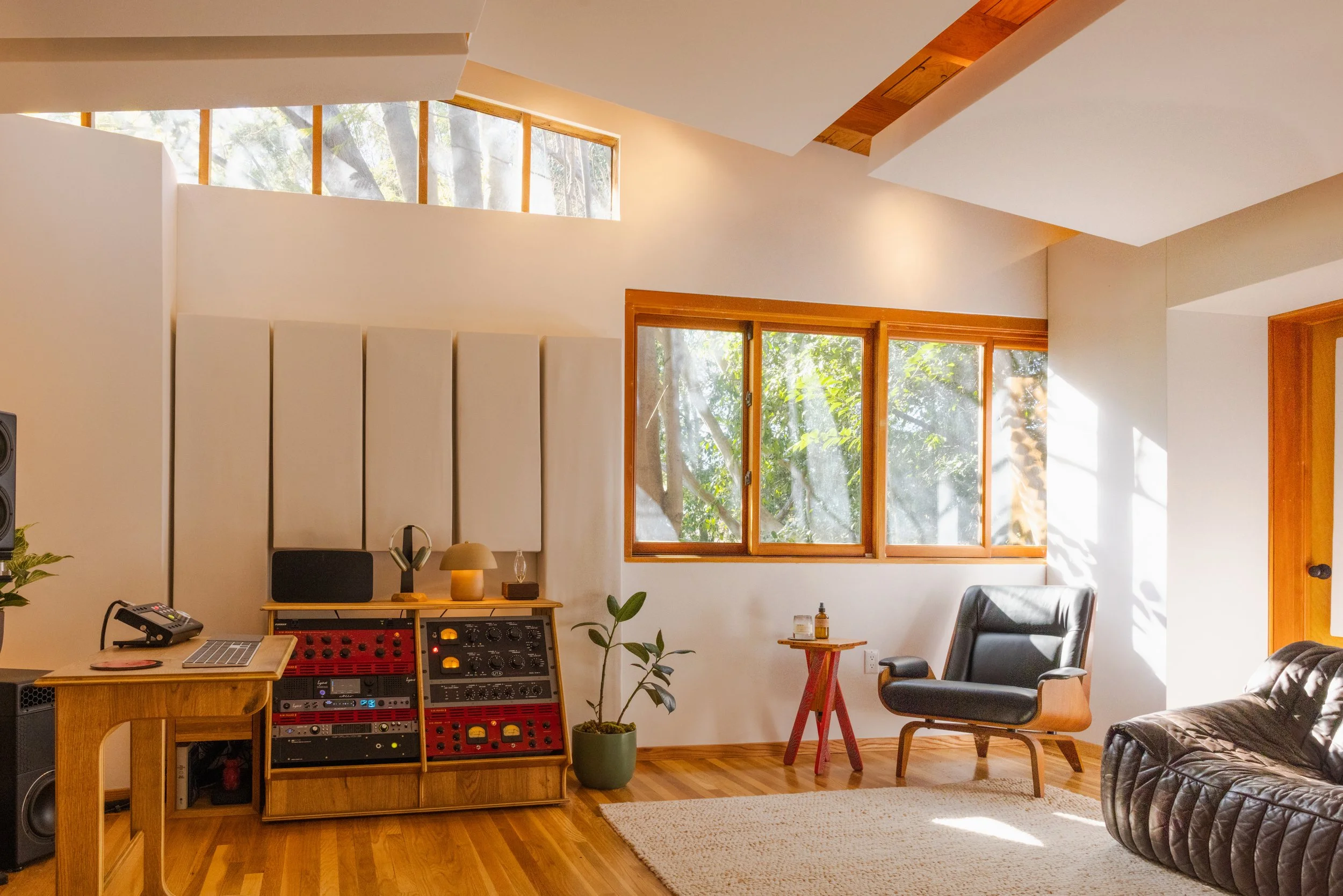 Interior of a modern living room with wooden floors, large windows, a black lounge chair, a leather couch, and a sound mixing console.