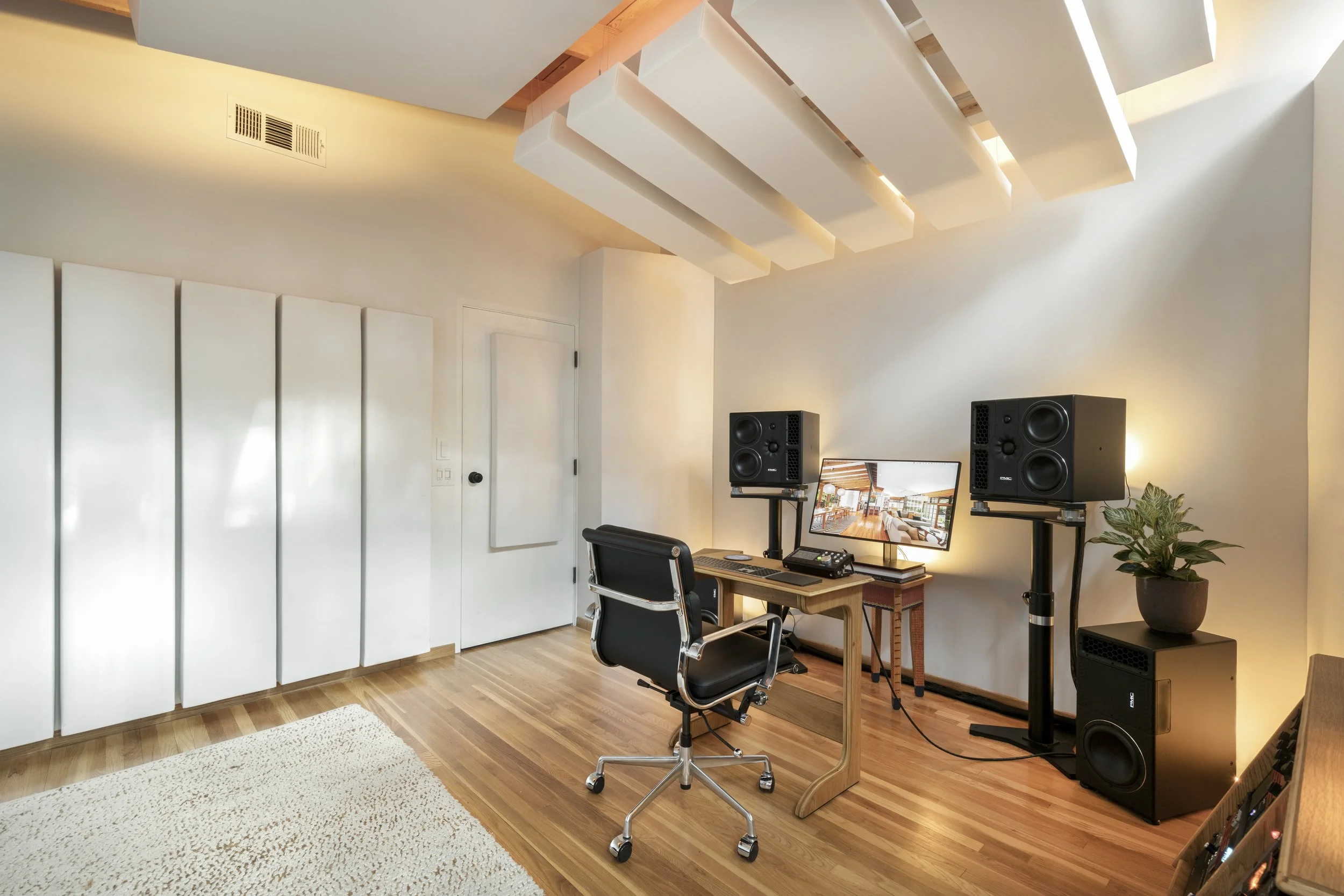 A modern home office with a wooden desk, black office chair, dual speakers on stands, a large monitor displaying a home interior, a plant, and wooden flooring with a beige rug.