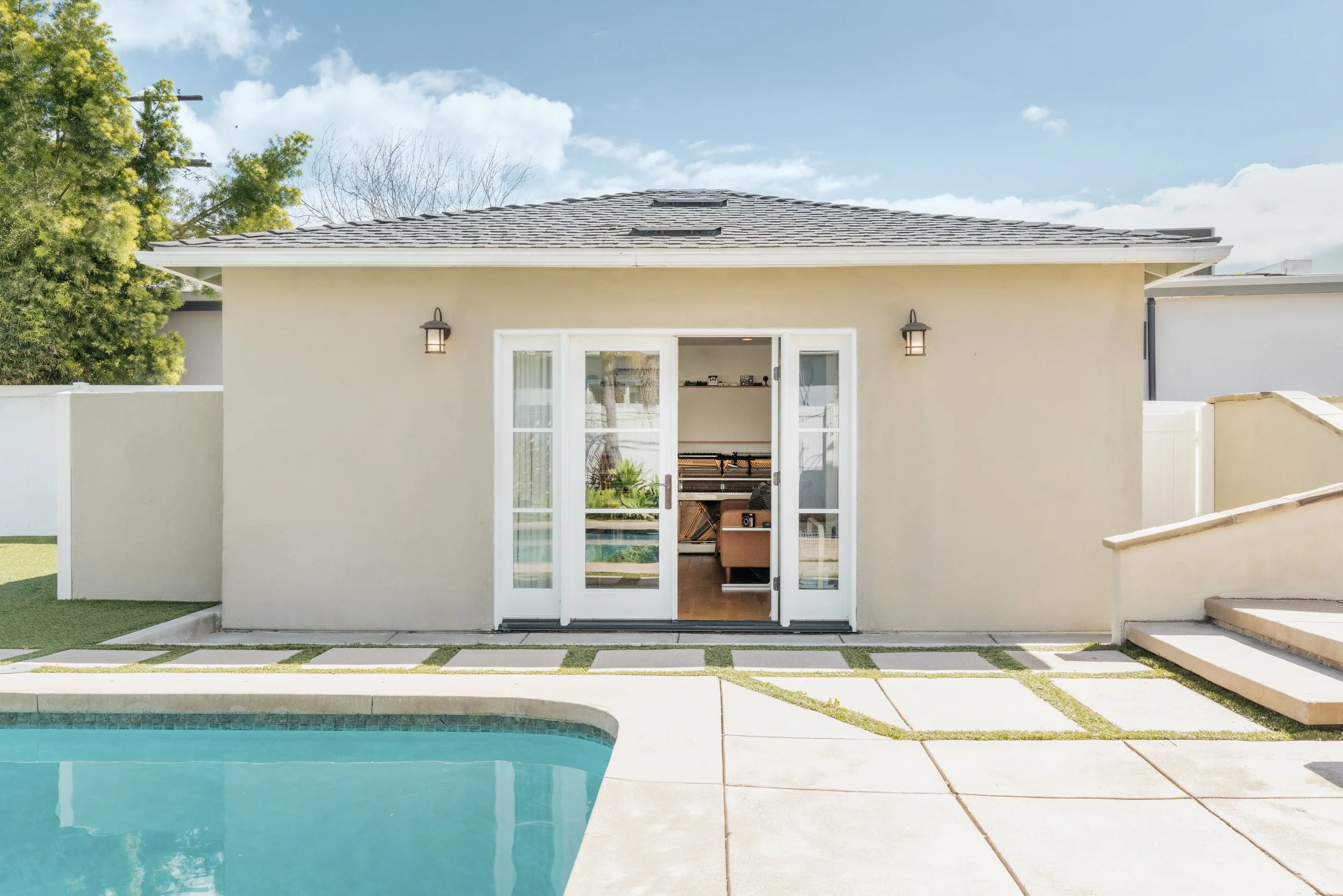View of the backyard pool area with a beige house in the background, open glass doors, and outdoor lighting fixtures.