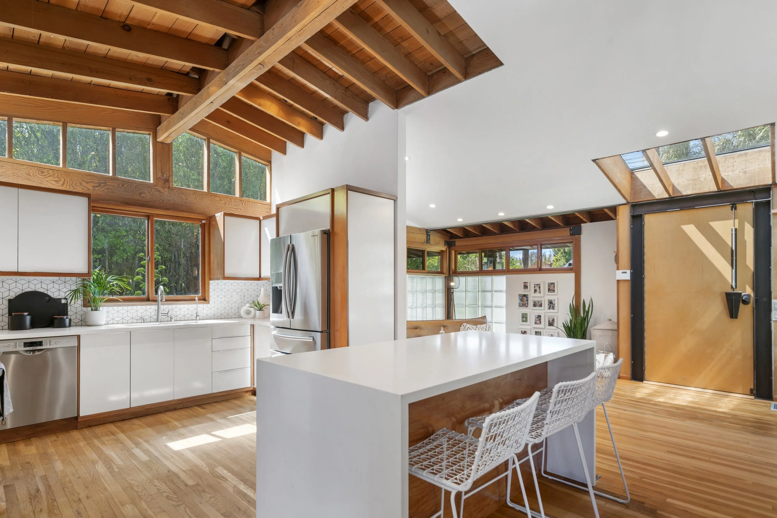 Modern kitchen with white cabinets, wooden ceiling beams, large windows, and a white island with wire chairs.