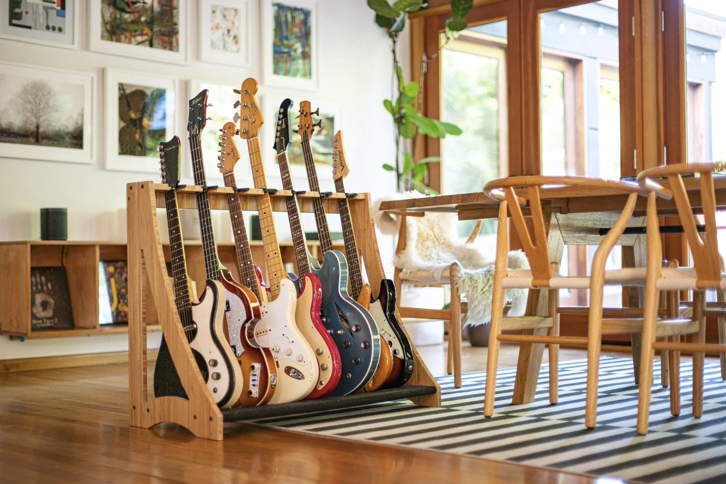 A collection of electric guitars on a wooden guitar rack in a bright, modern room with wooden furniture, framed artwork on the wall, and large windows letting in natural light.