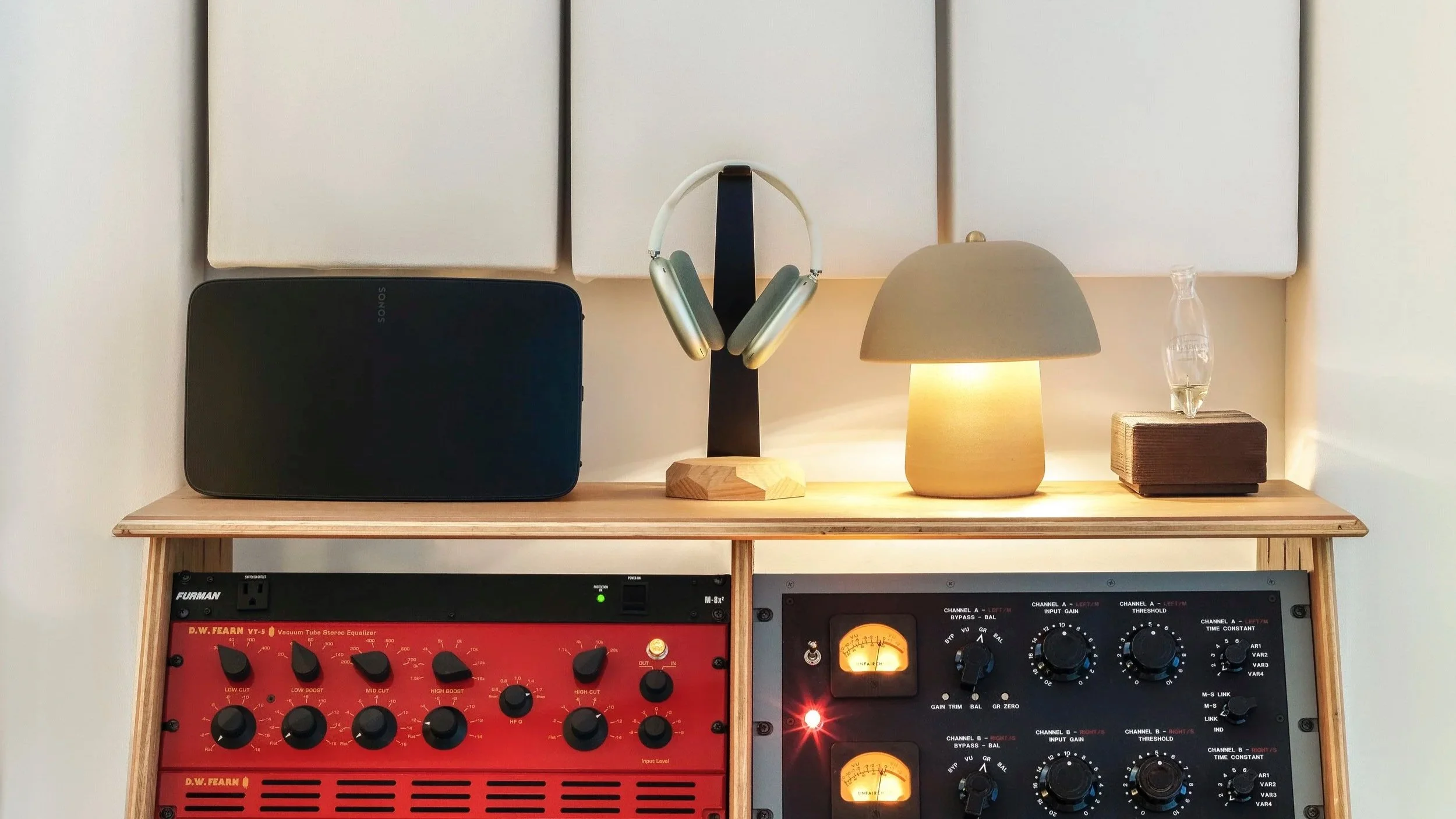 A music studio setup featuring a wooden console with audio equipment, a black Sonos speaker, a pair of headphones on a black and wood stand, a beige table lamp, and a small glass sculpture on a platform, with soundproof panels on the wall in the background.