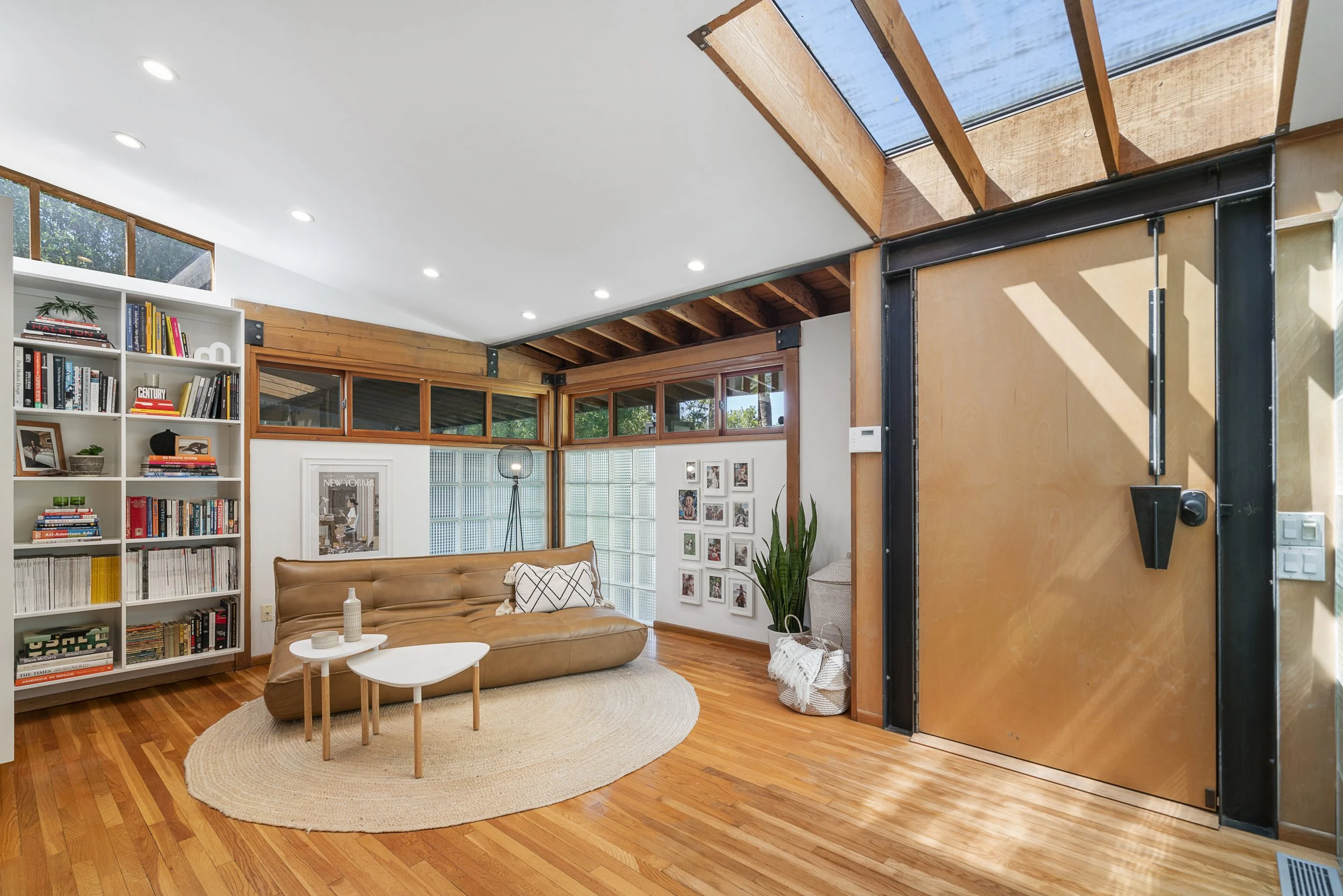 Modern living room with white walls, wooden accents, a leather sofa, a bookshelf filled with books, a gallery wall, and large windows letting in natural light.