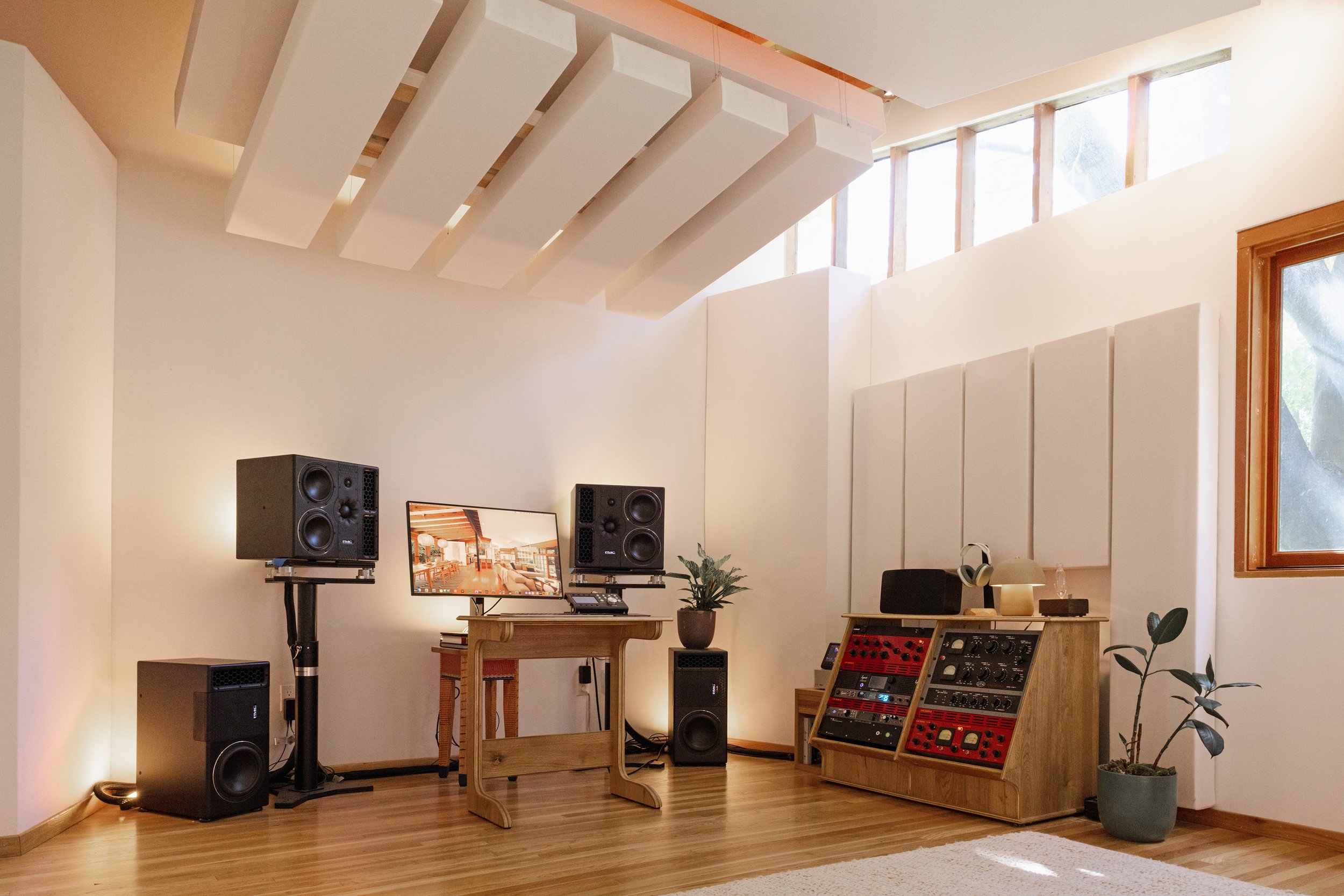 A sound studio with mixing equipment, speakers, a monitor, and plants in a room with wooden flooring and white walls.