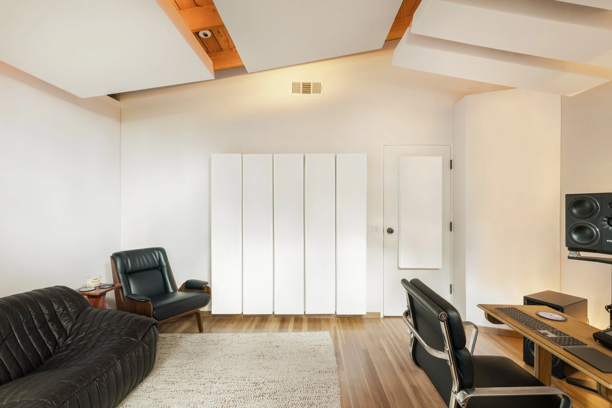 A modern recording studio or home office with white walls, wooden flooring, and acoustic panels on the ceiling. Contains a black leather sofa, a black armchair, a wooden desk with a keyboard, mouse, and a monitor, and a professional speaker in the corner.