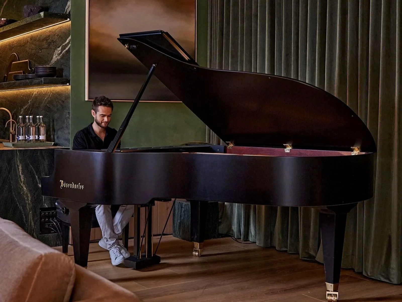 Man playing grand piano in a modern room with green curtains and dark marble walls.