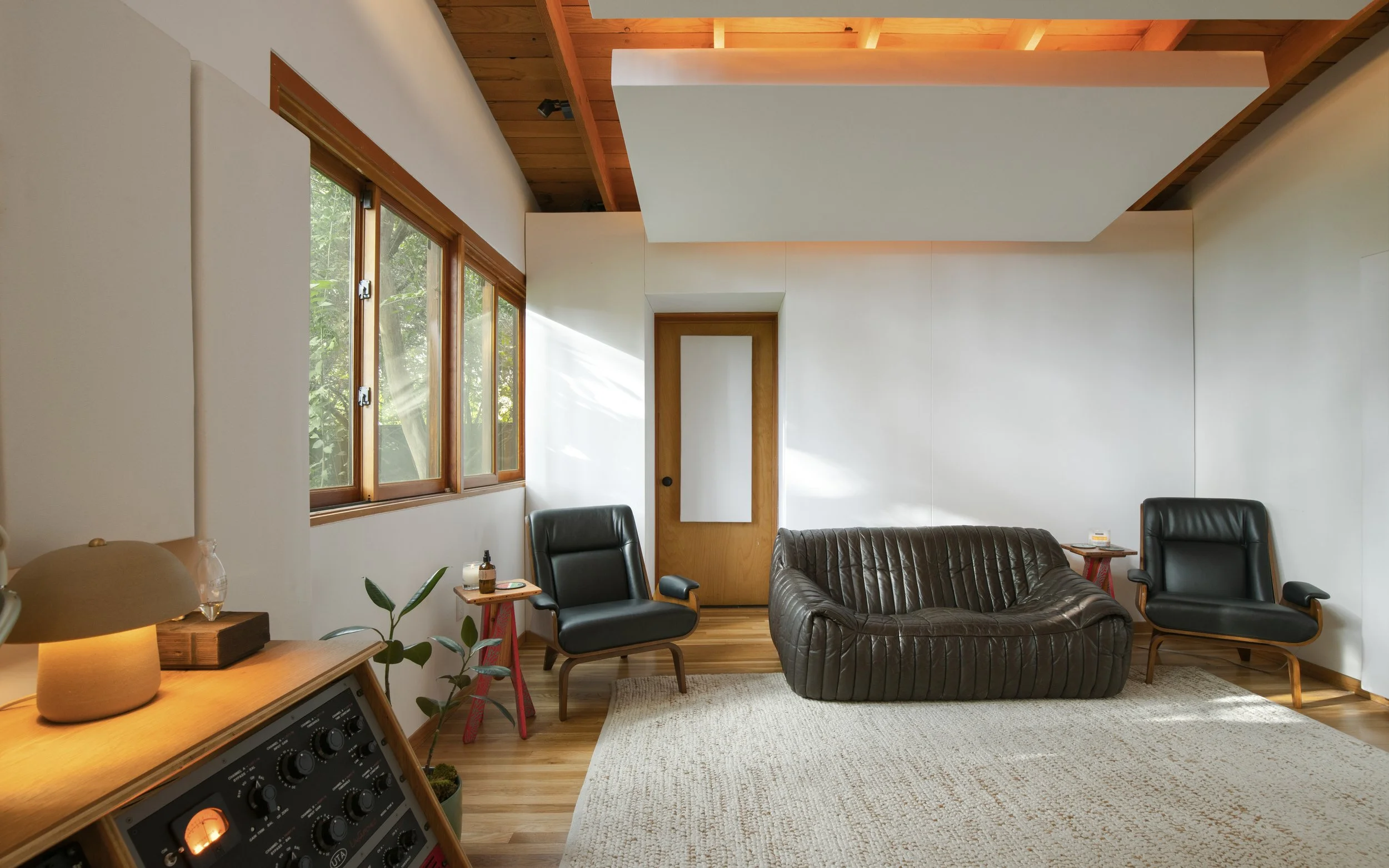A cozy living room with wooden ceiling and white walls. Contains a black leather armchair, a vintage brown leather sofa, and a black leather chair. Multiple small tables with decorative items and a potted plant. Large windows with a view of greenery outside and sunlight streaming in.