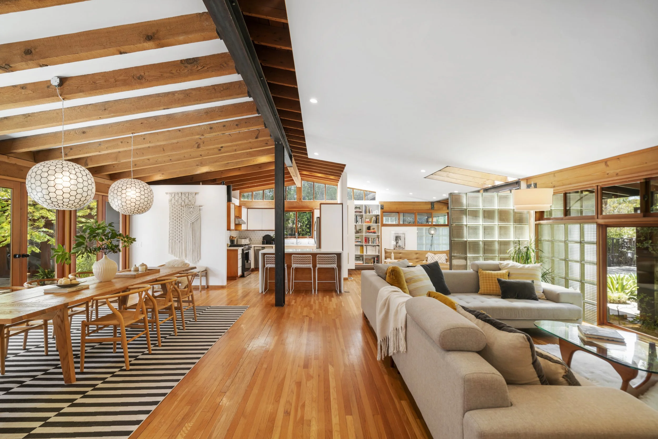Open-concept living and dining area with wooden ceiling beams, large windows, and modern decor.