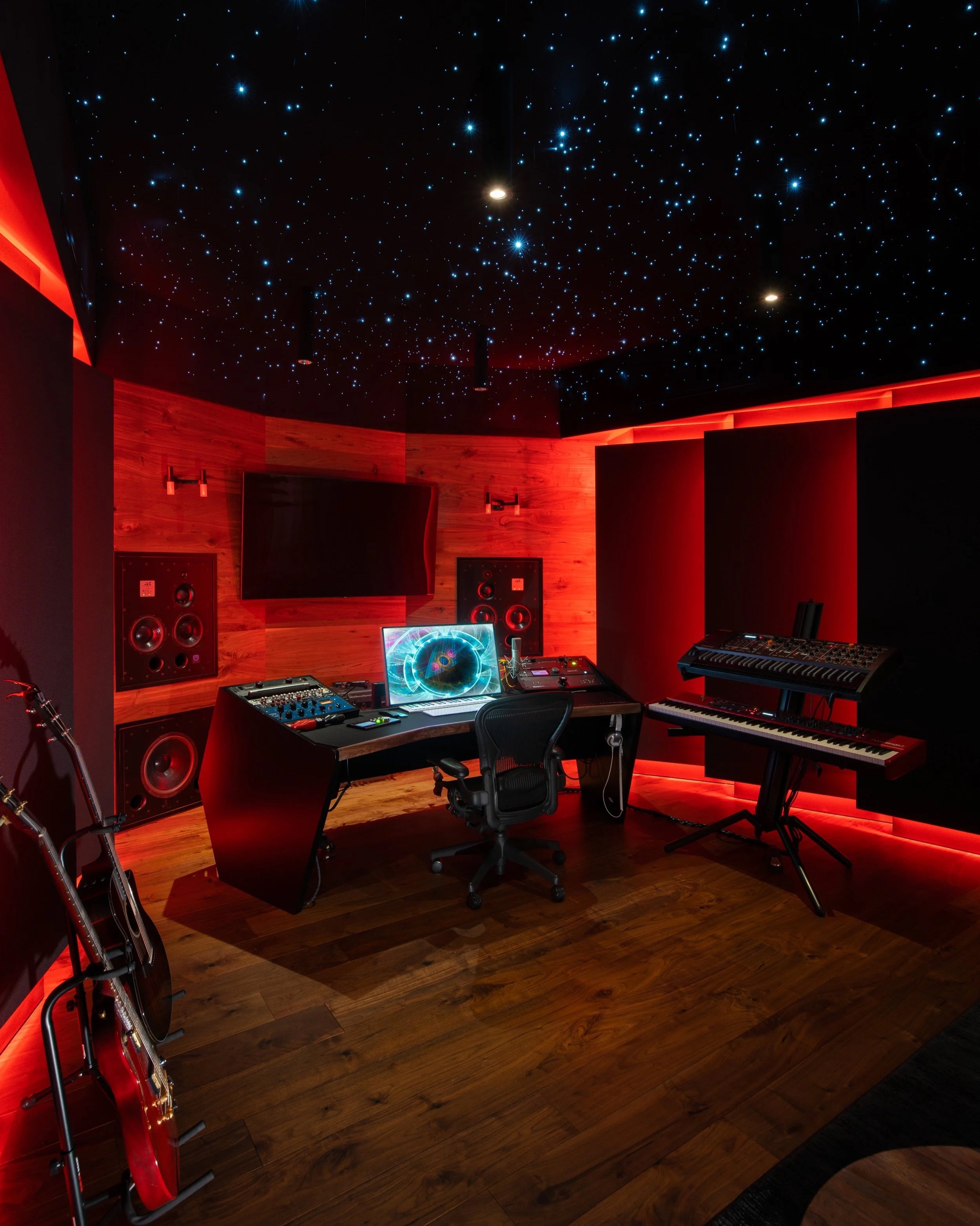 Music recording studio with wooden walls, starry sky ceiling, large black monitor, computer with glowing cosmic wallpaper, speakers, mixing console, keyboard, two guitars, and red ambient lighting.