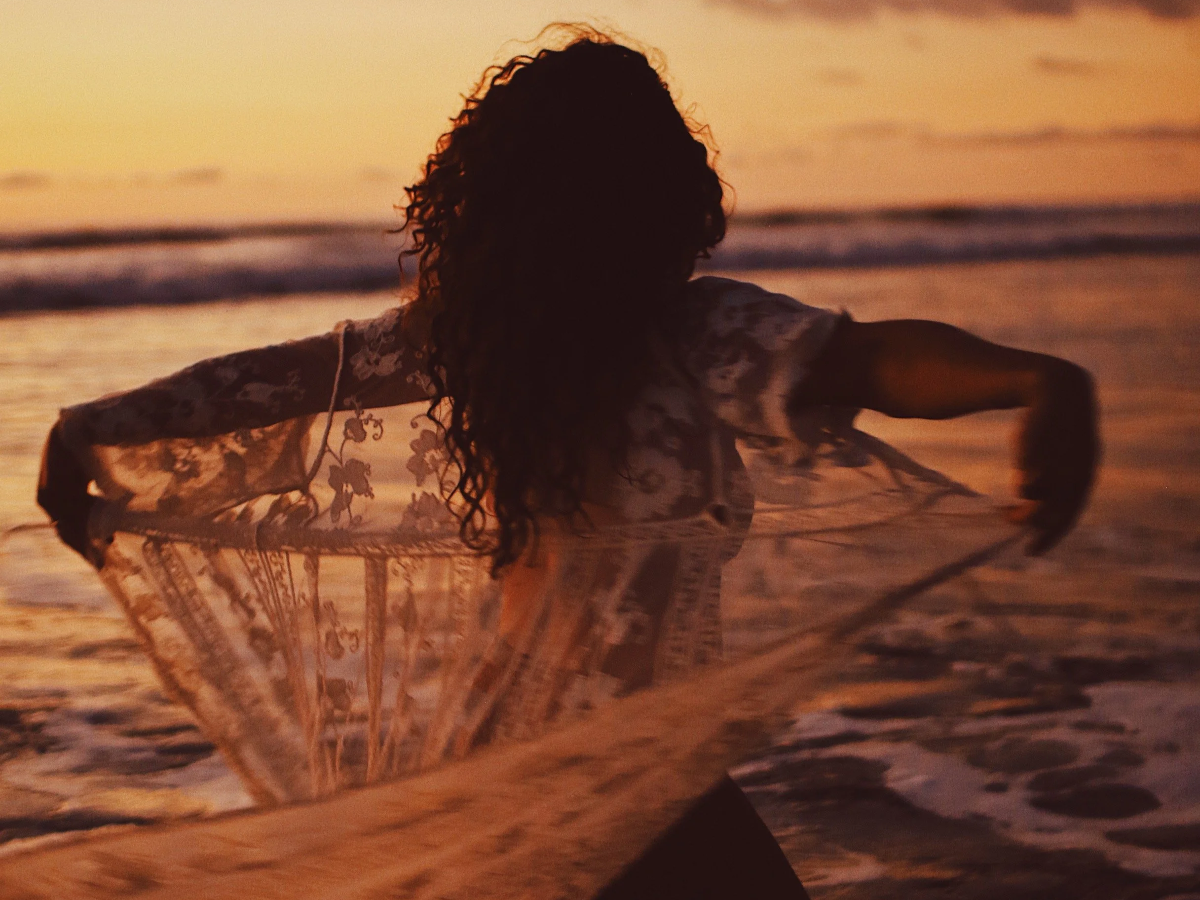 A person with curly hair wearing a sheer floral shawl or wrap, standing on a beach during sunset, with their arms outstretched.