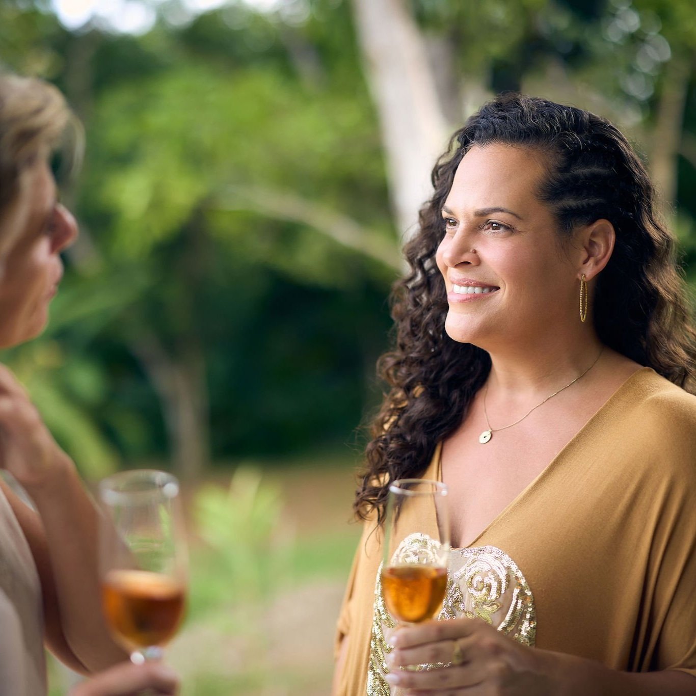 A woman with dark curly hair and gold jewelry smiling while holding a glass of champagne during an outdoor gathering.