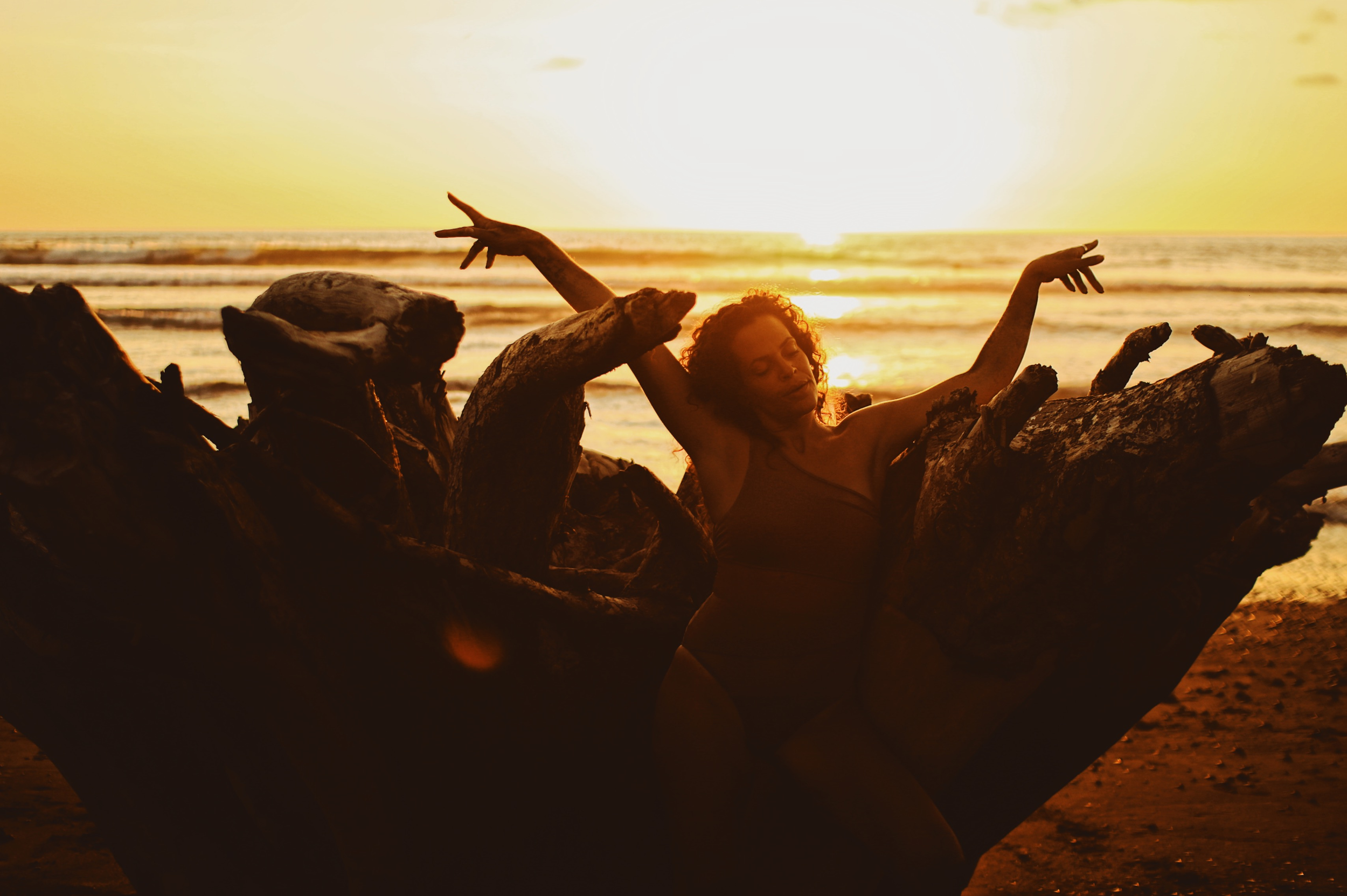 A woman with curly hair sits among driftwood on a beach at sunset, with the ocean and sun in the background.