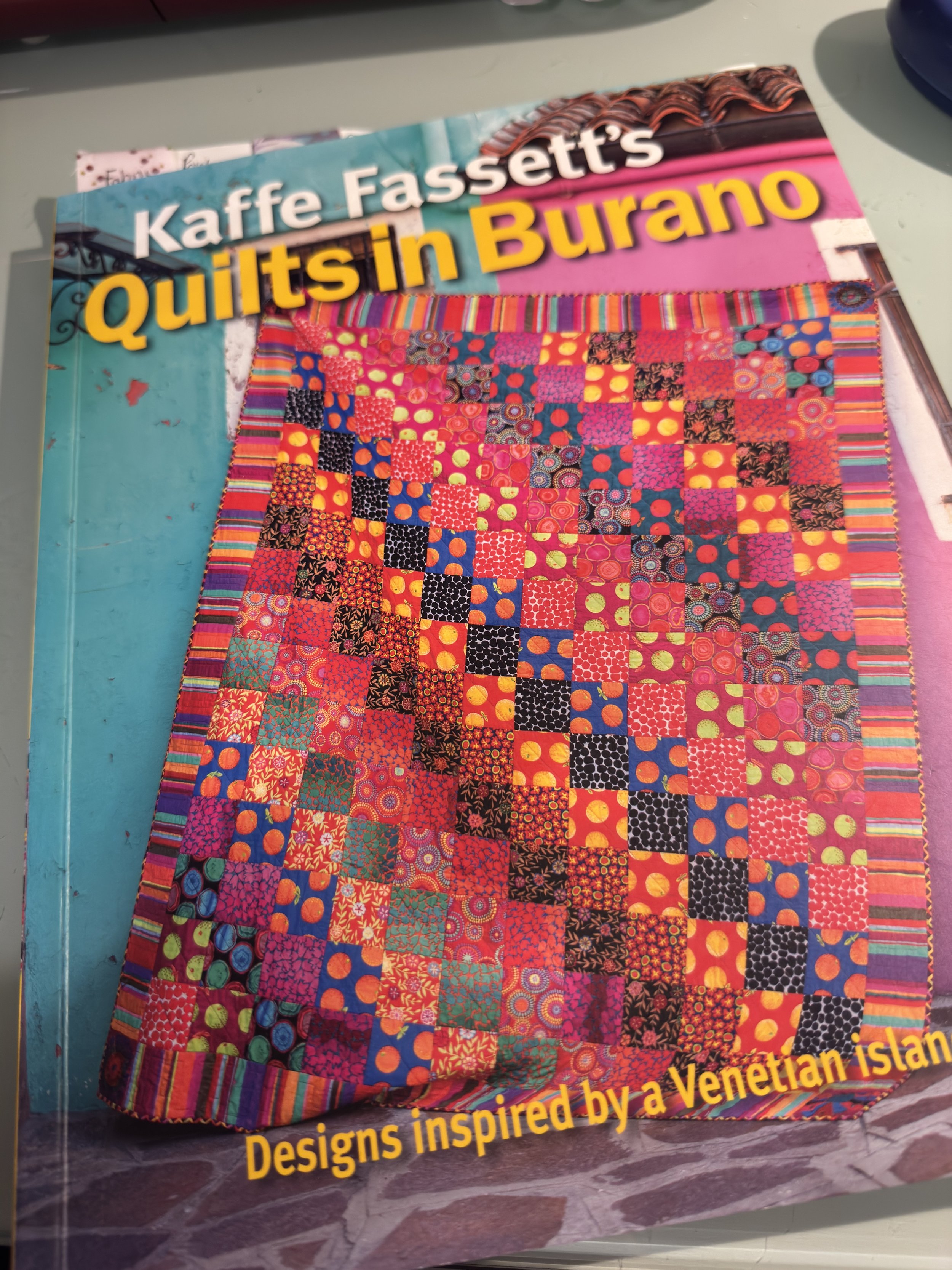 Front cover of the quilting book "Kaffe Fassett's Quilts in Burano" featuring a vibrant, multi-colored patchwork quilt inspired by Venetian island designs.