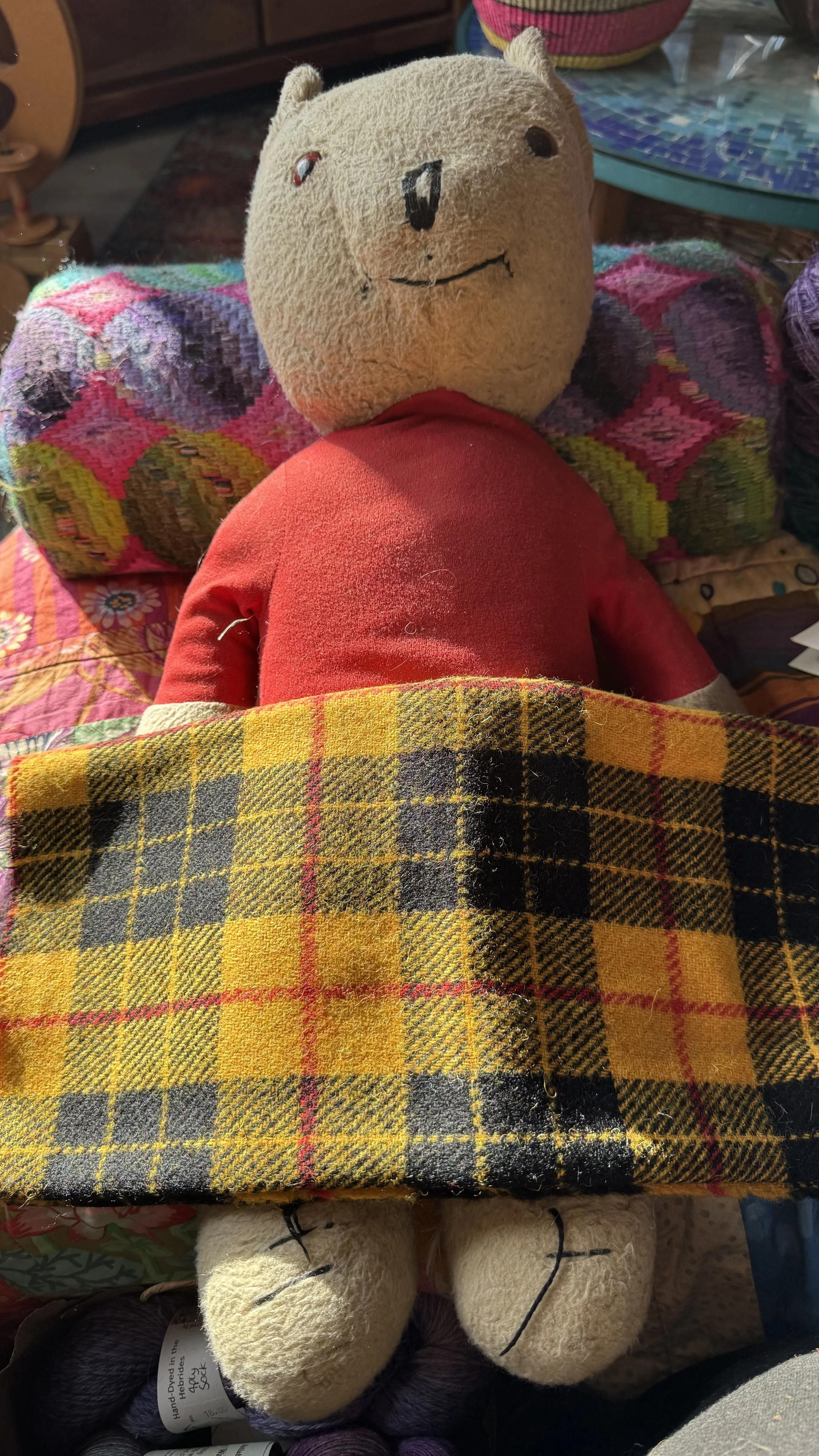 he vintage Rupert the Bear toy nestled under a piece of vibrant yellow, black, and red checked Harris Tweed fabric, with colorful patchwork cushions visible in the background.
