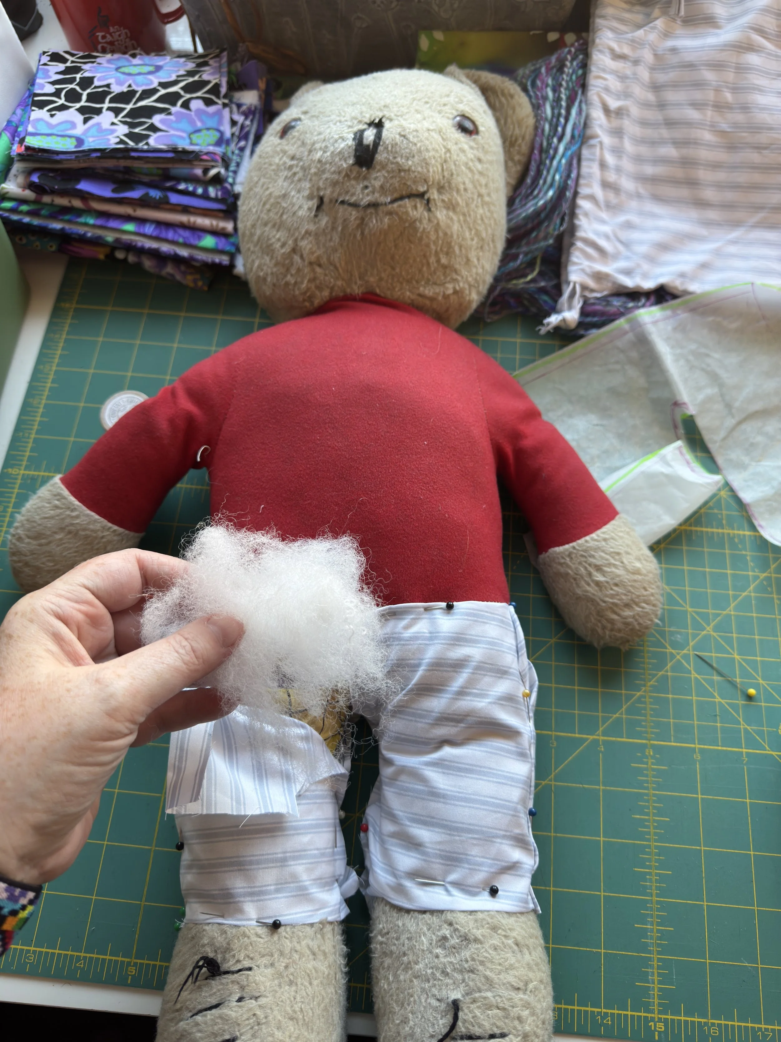 A top-down view of the vintage Rupert the Bear toy on a green cutting mat during a repair. A hand holds a tuft of white stuffing over the bear's legs, which are pinned into new white and blue striped cotton "long john" liners.