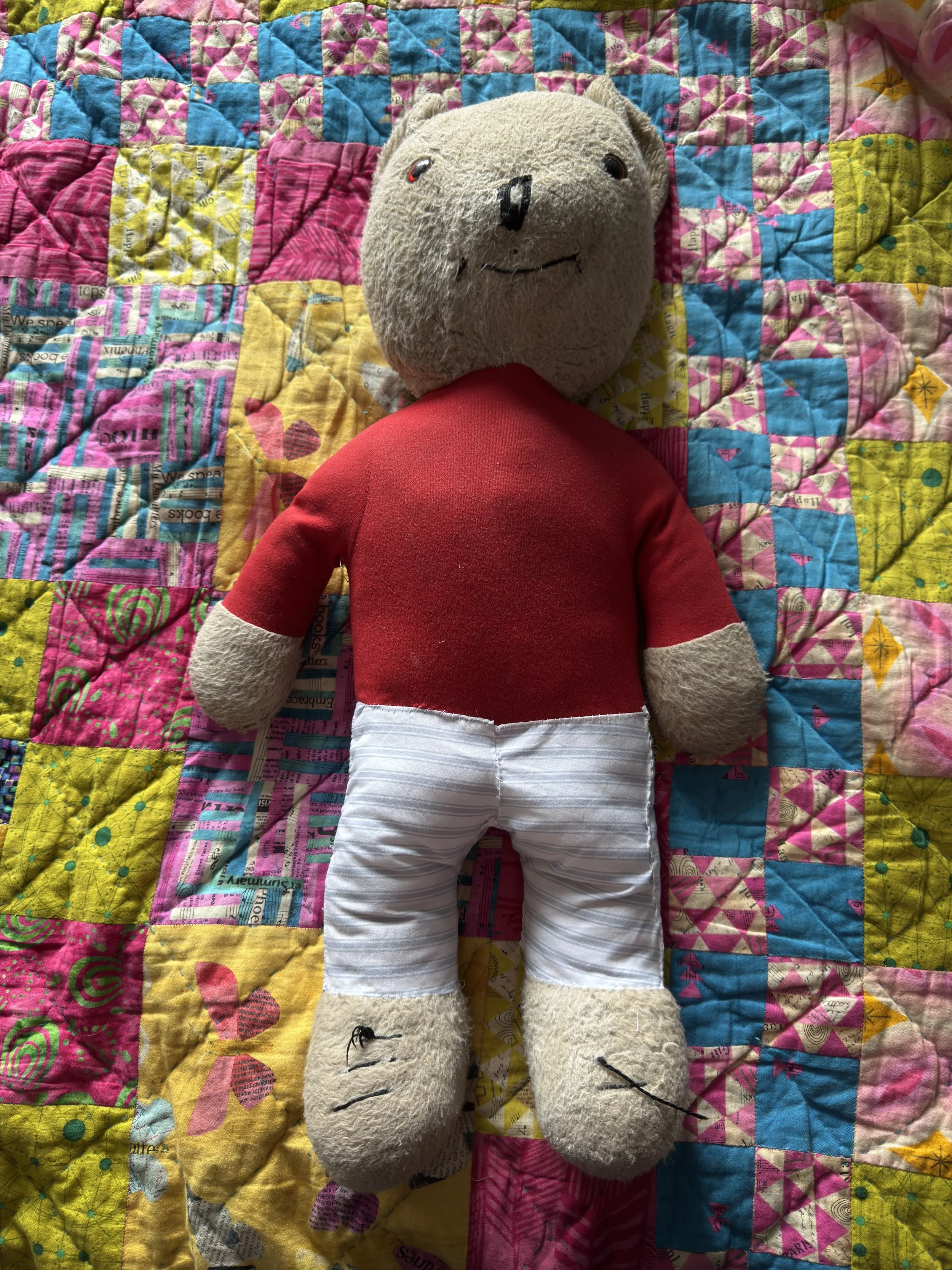 he vintage Rupert the Bear toy lying on a colorful, vibrant patchwork quilt. The bear is wearing his red sweater and the newly finished white and blue striped cotton undergarment liners, showing his restored shape before his final trousers are added