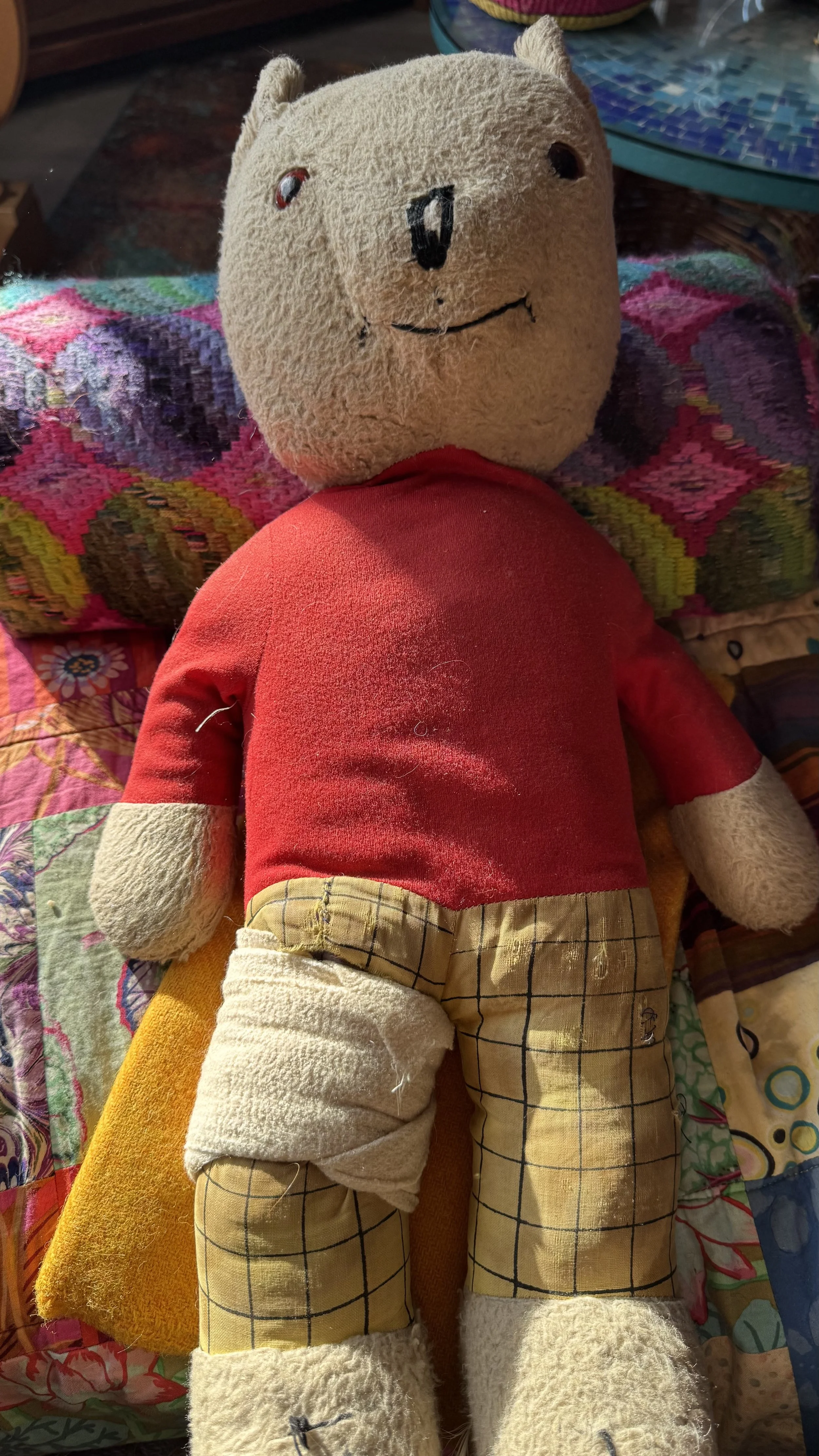 A close-up of a vintage Rupert the Bear toy sitting on a colorful patchwork quilt. The bear has a worn, cream-colored face, a red sweater, and yellow checked trousers. One leg is wrapped in a thick white fabric bandage to hold in stuffing.