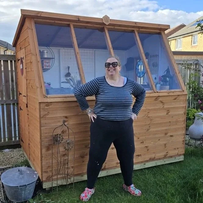 A smiling woman standing in front of a new wooden garden craft shed with large front windows.