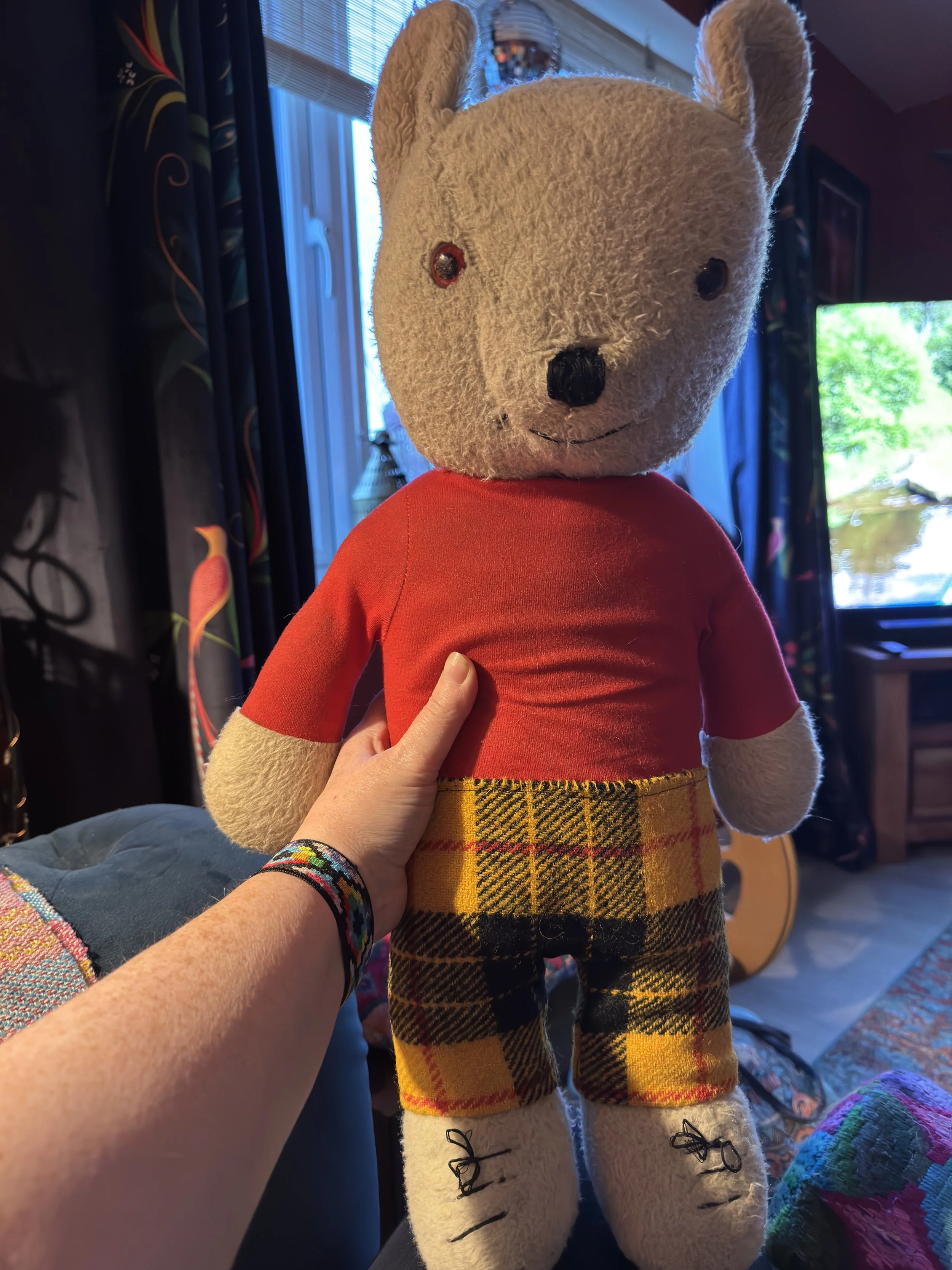 The restored vintage Rupert the Bear toy standing upright, featuring his original red sweater and newly crafted yellow and black checked Harris Tweed trousers with a red pinstripe.