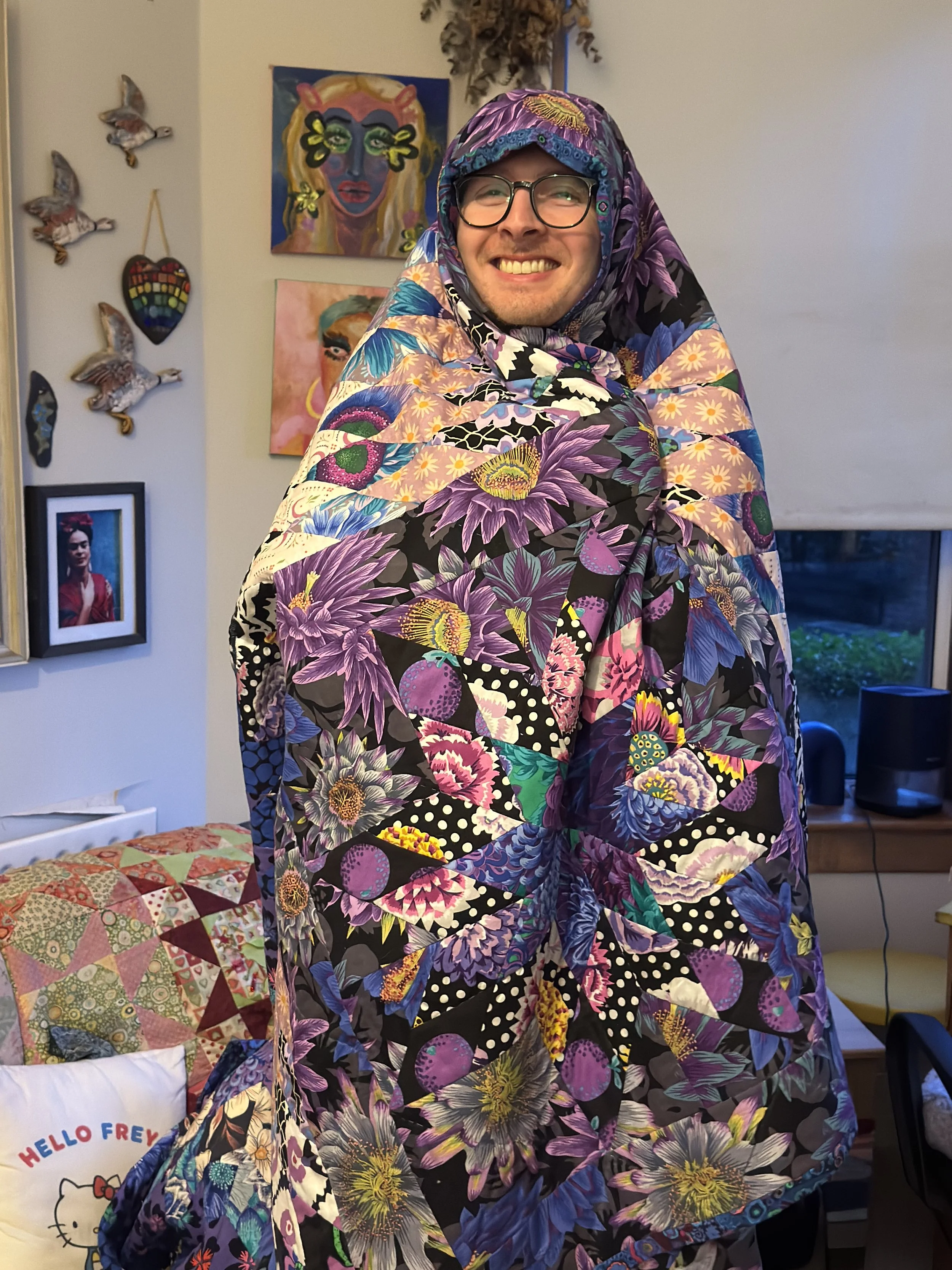 A man cocooned in a finished Shimmer Star patchwork quilt in shades of purple, blue, and green.
