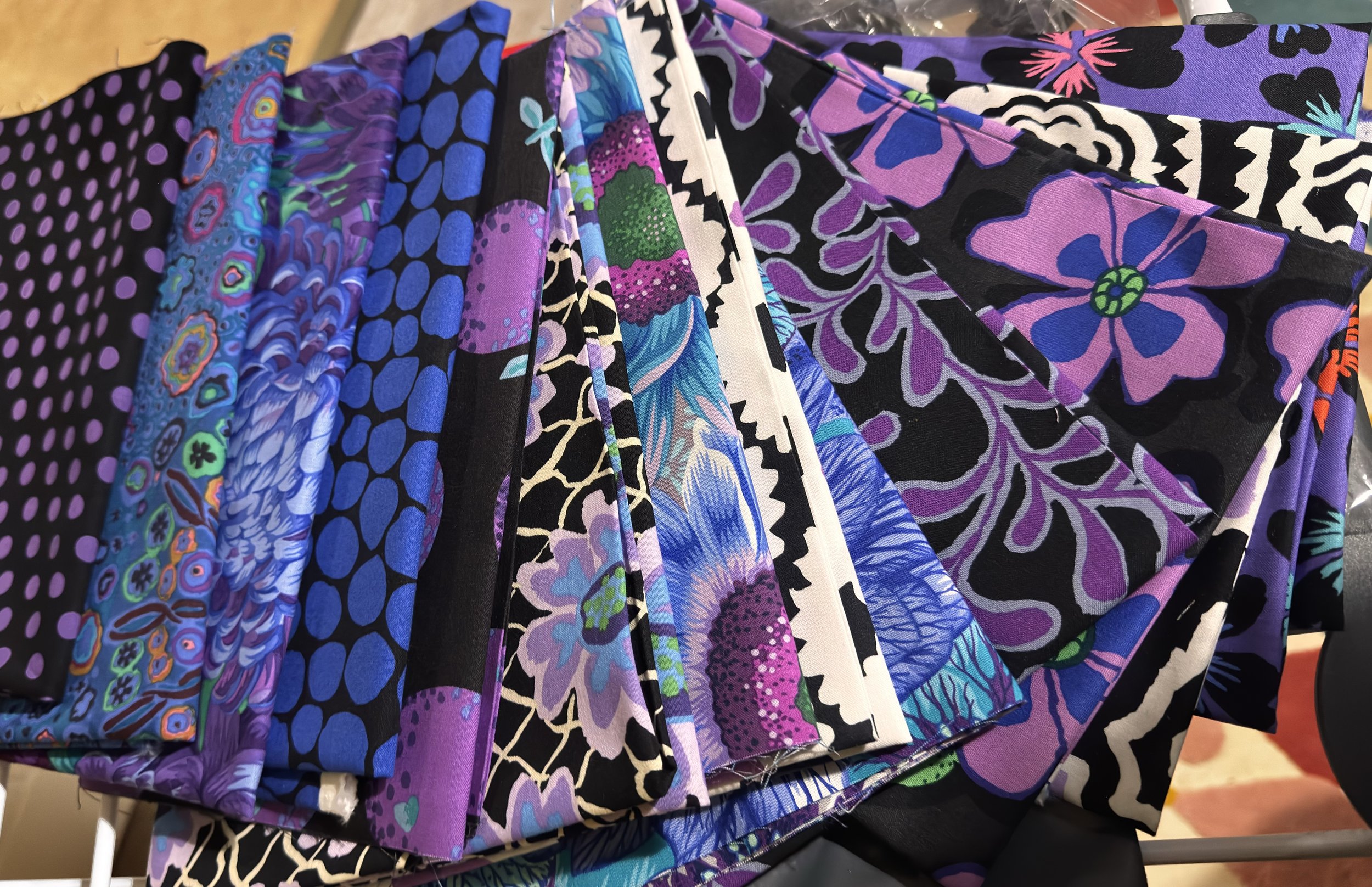 A fanned-out pile of folded fabric  in various purple, blue, black, and floral patterns, ready for patchwork piecing.