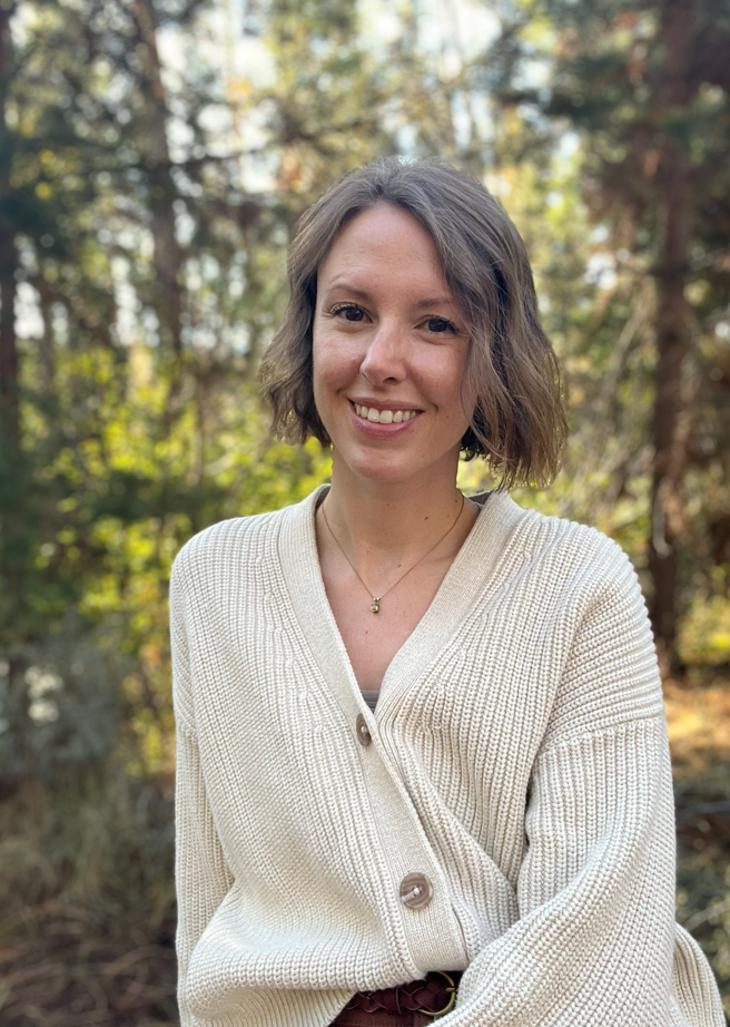 Lauren, owner of Rivers And Roads Counseling with short brown hair and a light complexion, smiling in an outdoor setting with trees and sunlight in the background, wearing a cream-colored cardigan.