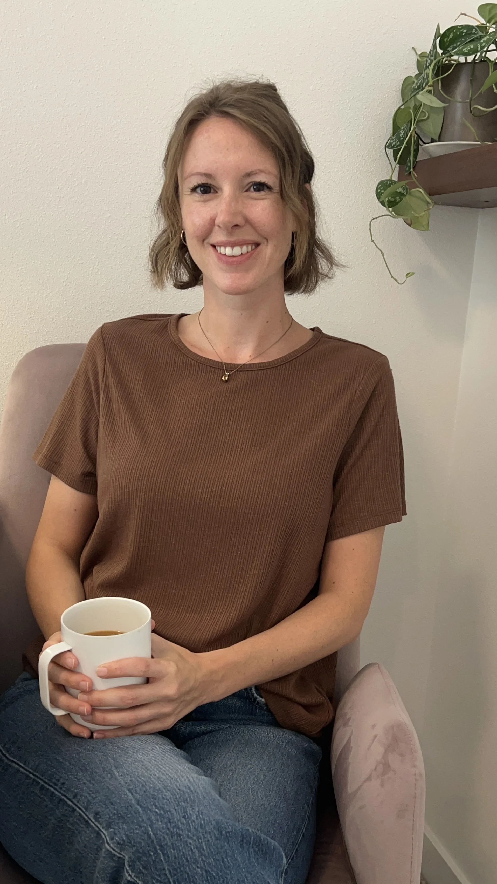 Lauren, owner of Rivers And Roads Counseling - with shoulder-length wavy hair, wearing a brown T-shirt and jeans, sitting on a chair with a cup of coffee in her hands, smiling at the camera. There is a potted plant on a wall shelf in the background.