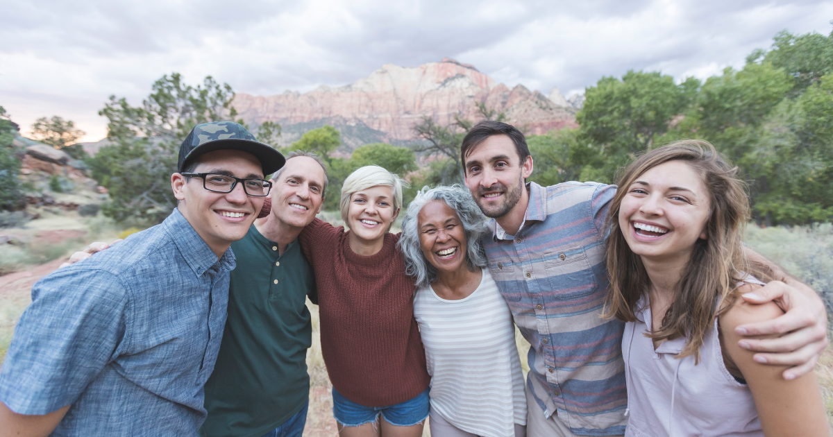 Group of six diverse people smiling and embracing outdoors with mountains and trees in the background.