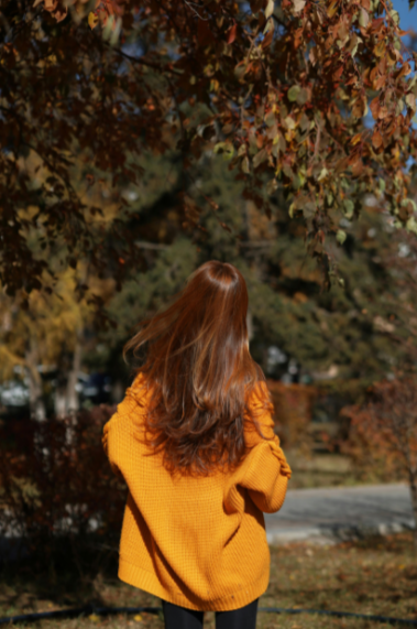 A woman with long red hair wearing a yellow sweater standing outdoors in front of autumn trees.