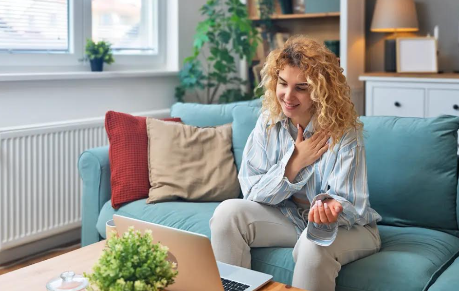 A woman with curly hair sitting on a light blue sofa, holding her chest and smiling while looking at her laptop on the coffee table, in a cozy living room.