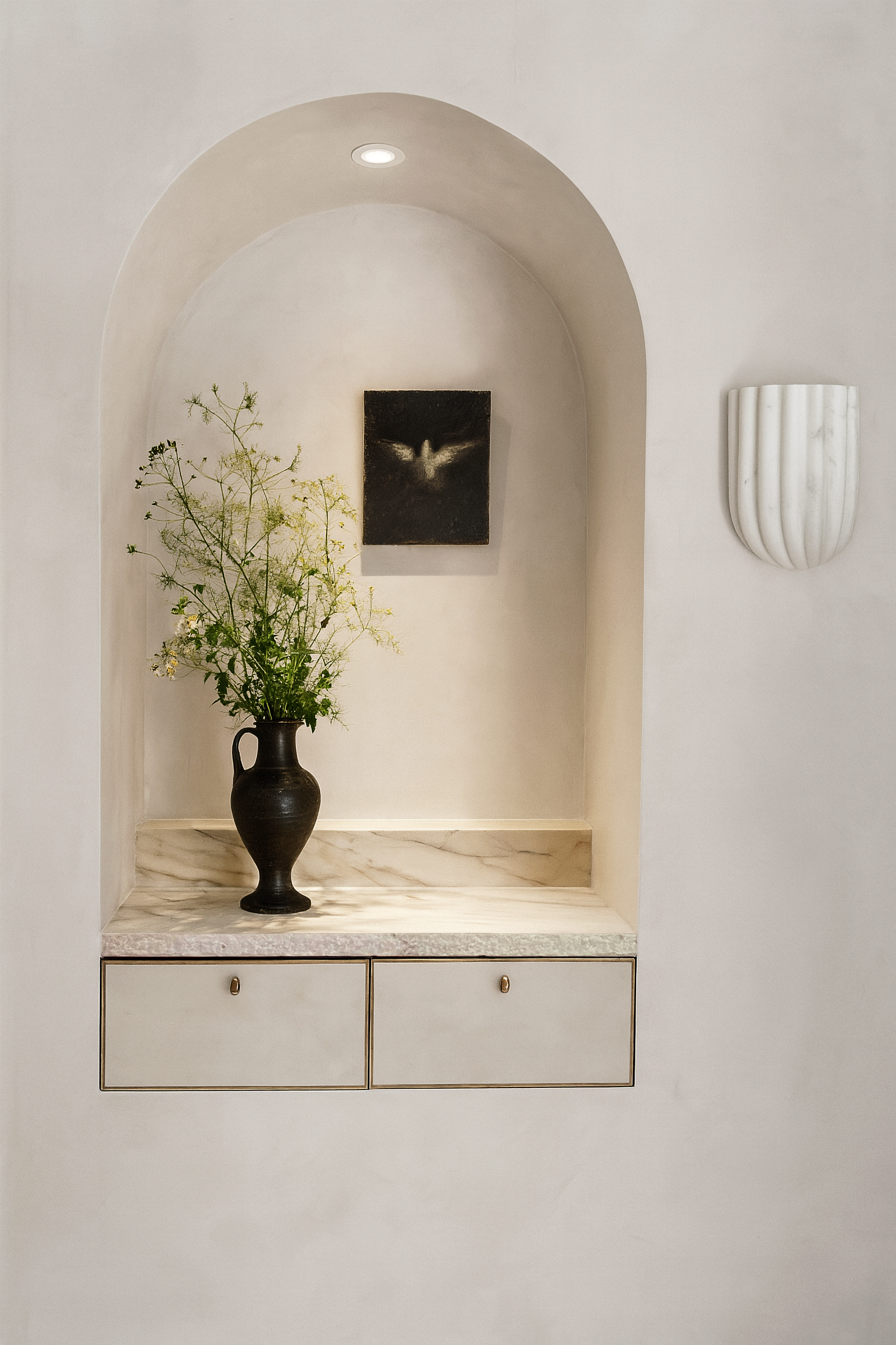 A minimalist interior niche with a marble surface, decorated with a black vase containing tall green and white flowers, a small black painting, a modern white marble wall sconce, and a white cabinet with gold accents.